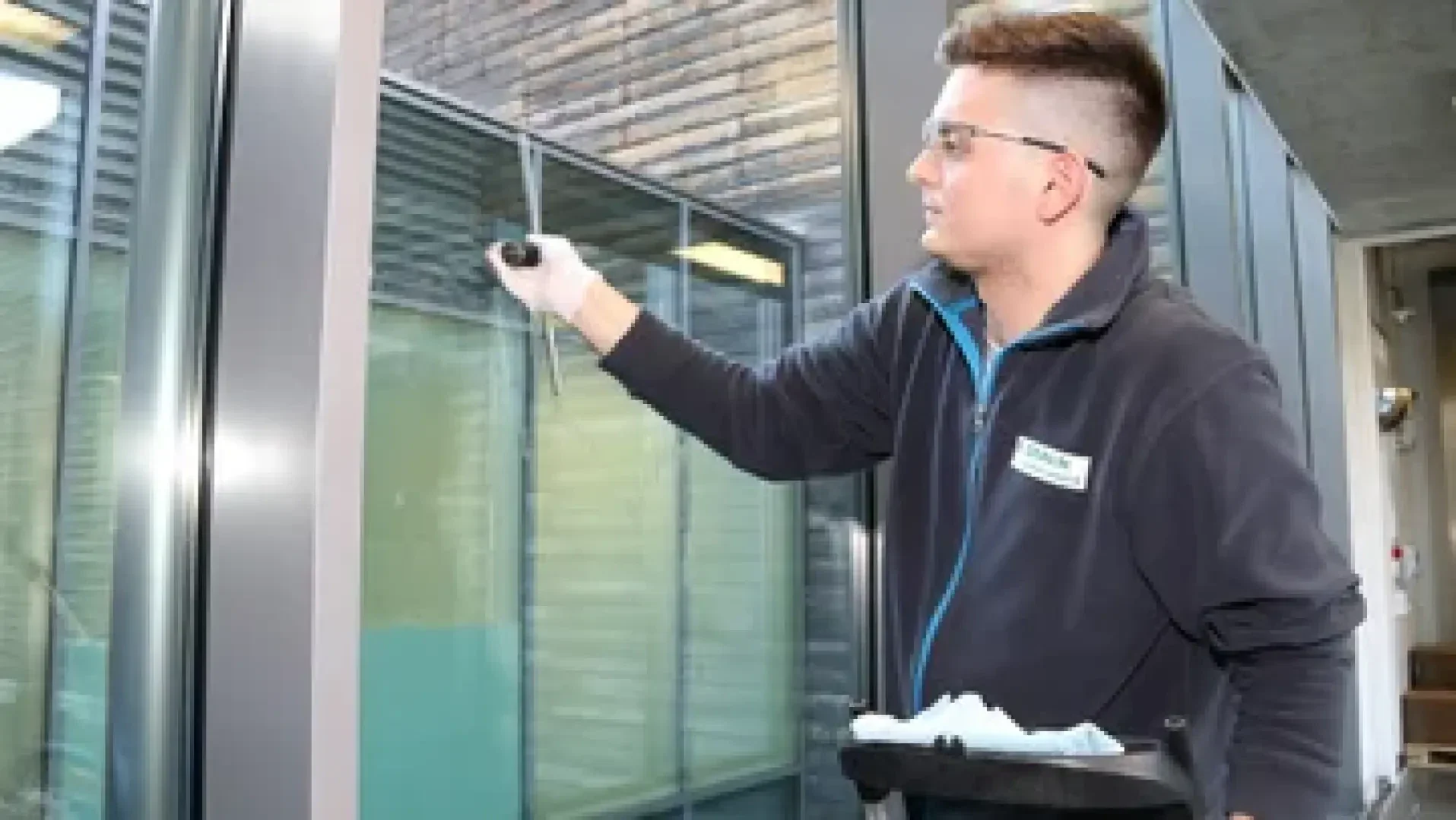 Ein Mann mit Schutzbrille reinigt ein großes Fenster mit einem Abzieher. Er steht drinnen in der Nähe eines Metallrahmens, neben ihm steht ein Wagen mit Reinigungsutensilien.