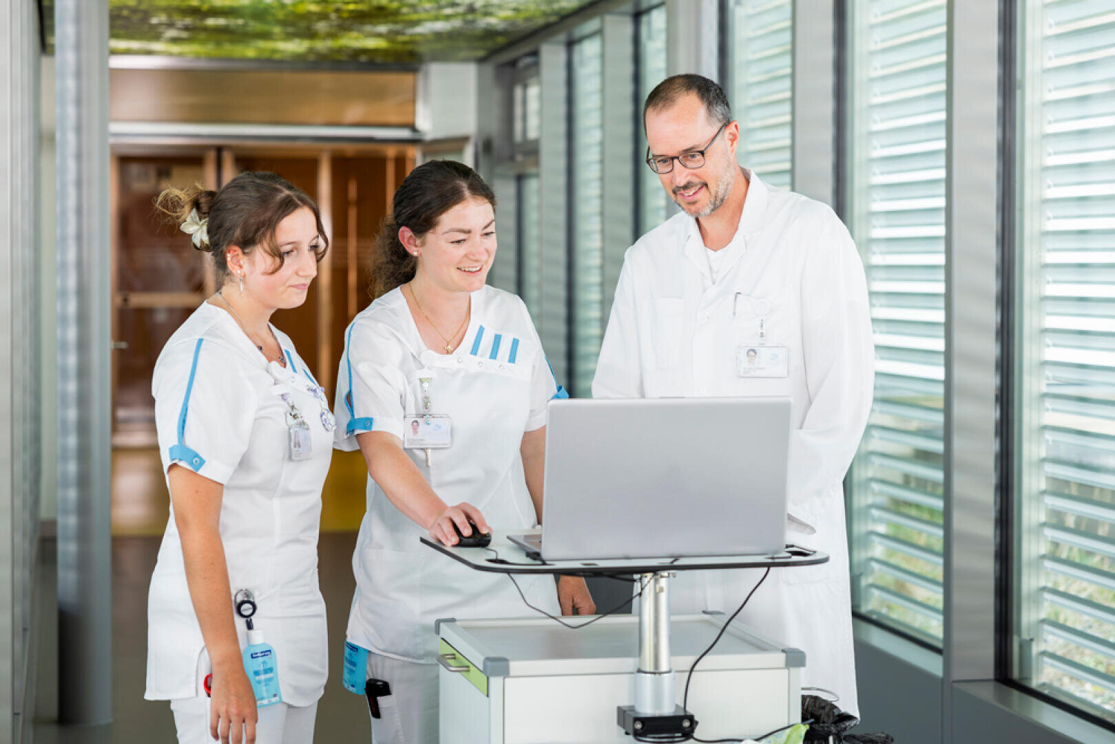Drei medizinische Fachkräfte in weißen Uniformen stehen in einer modernen medizinischen Einrichtung mit Glaswänden neben einem Computer auf einem Rollwagen, lächeln und blicken auf den Bildschirm.