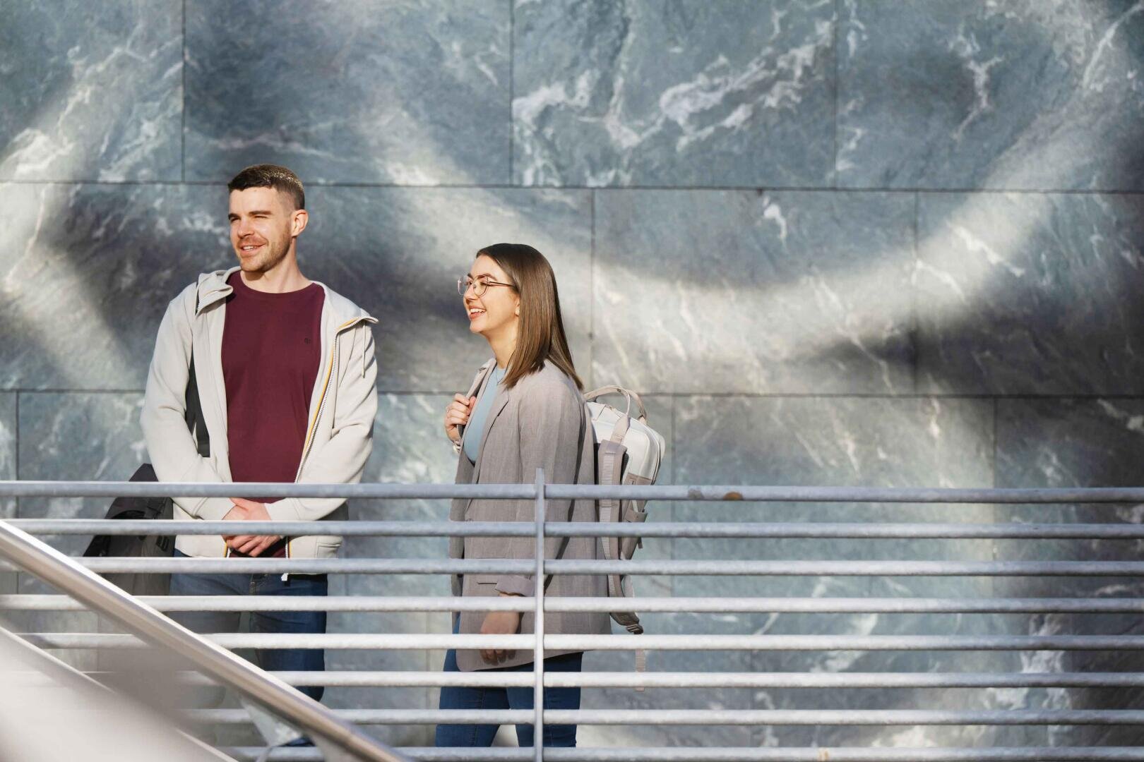 Zwei Personen mit Rucksäcken stehen lächelnd an einem Metallgeländer vor einer großen, strukturierten grauen Marmorwand mit kreisförmigen Lichtmustern.