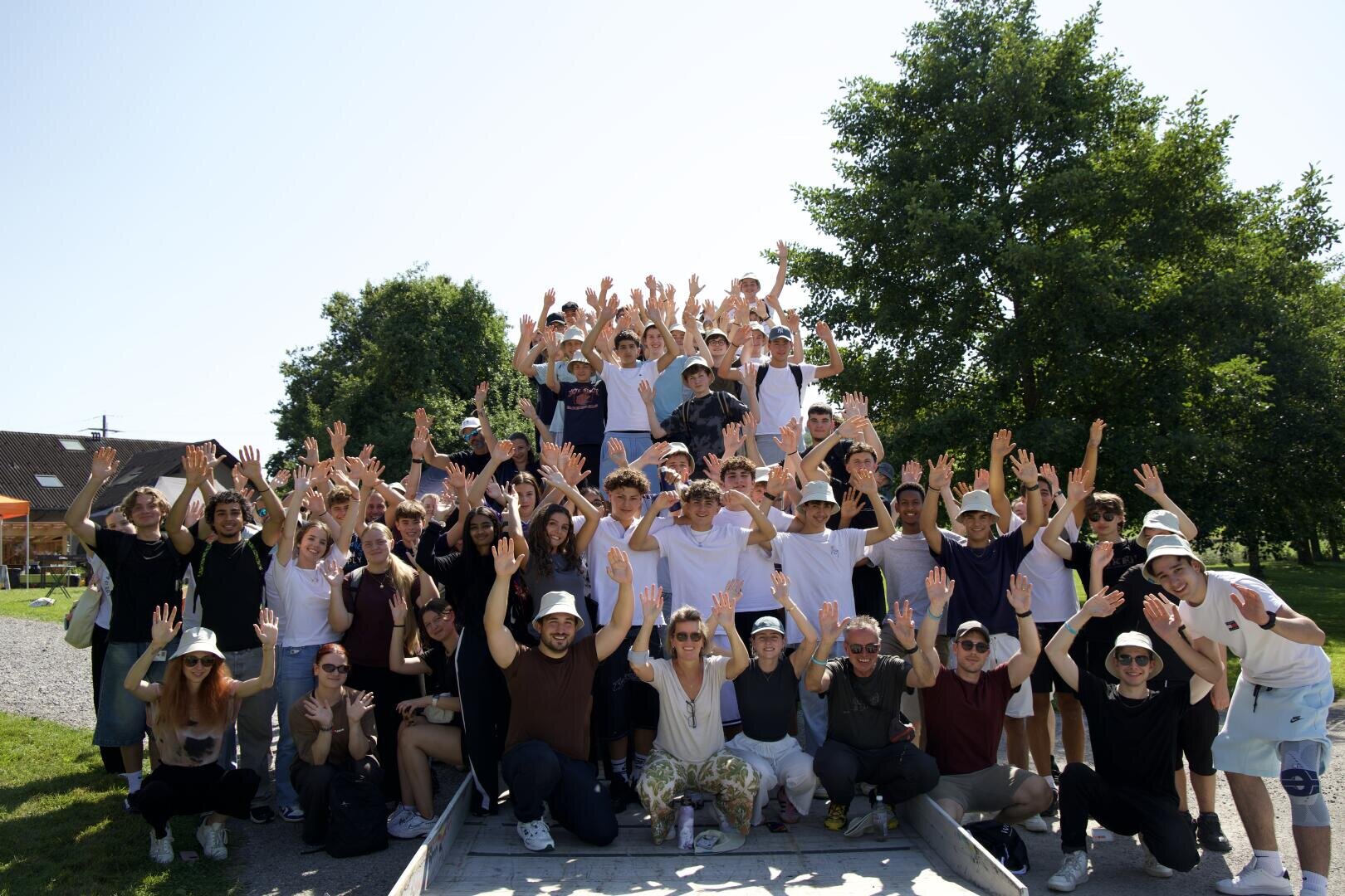 Eine große Gruppe von Menschen, meist junge Erwachsene, posiert an einem sonnigen Tag im Freien, lächelt und hebt die Hände. Sie stehen auf Stufen, umgeben von grünen Bäumen und Gras. Alle wirken glücklich und voller Energie.
