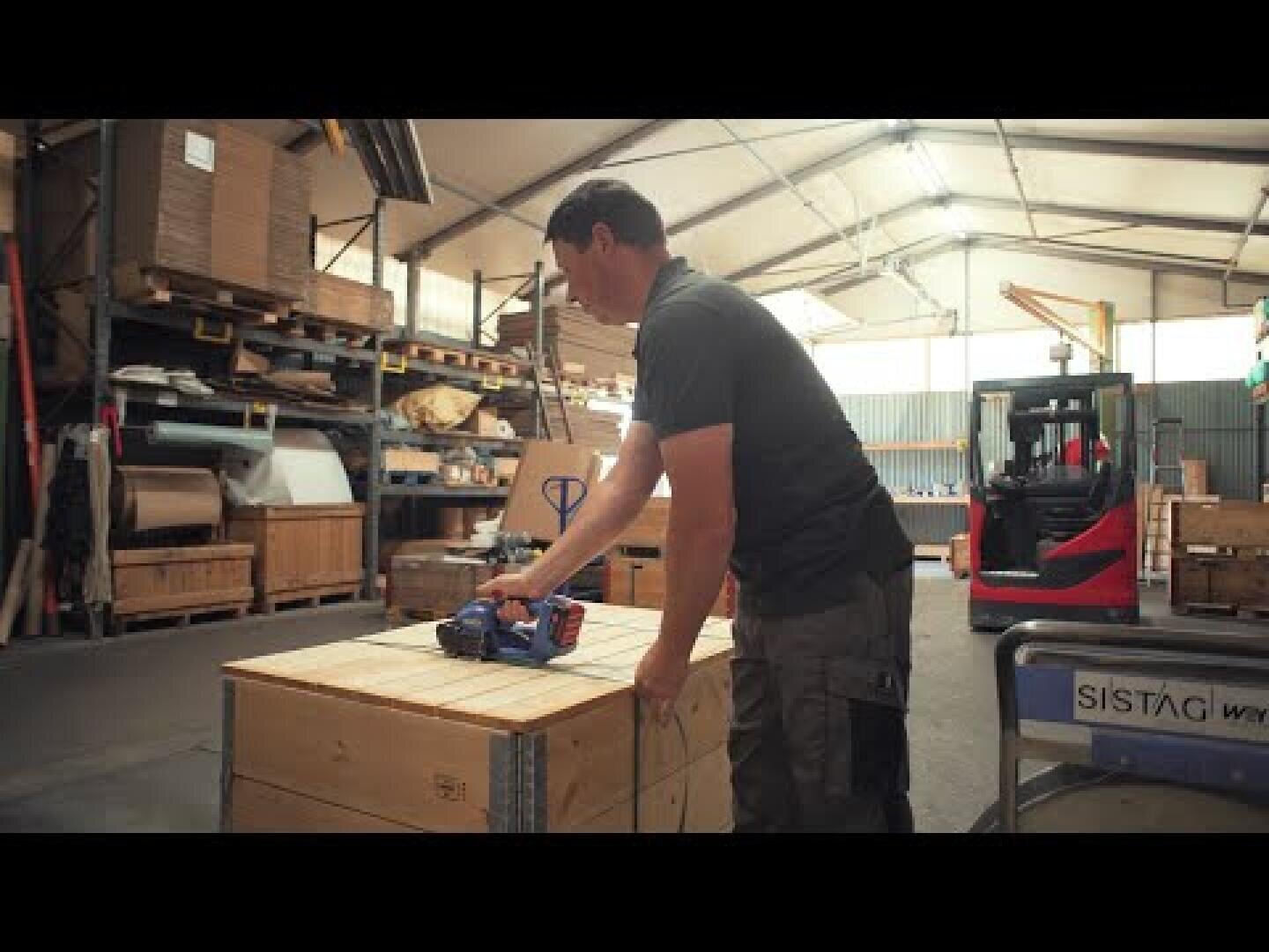 Ein Mann steht in einem Lagerhaus und arbeitet mit einem handgeführten Werkzeug an einer Holzkiste. Im Hintergrund sind Regale mit Kisten und ein roter Gabelstapler zu sehen. Der Raum ist gut beleuchtet und hat hohe Decken.