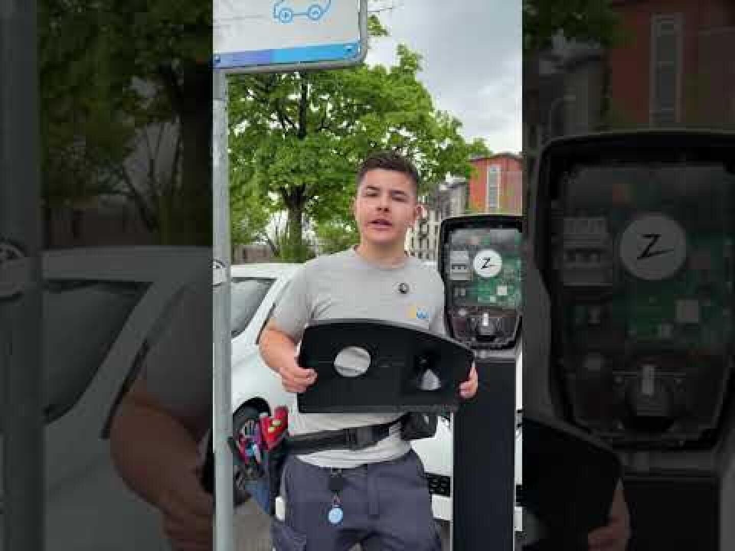 Ein Arbeiter steht vor einer Ladestation für Elektrofahrzeuge und hält die vordere Abdeckung der Ladestation. Im Hintergrund sind ein weißes Auto und Bäume zu sehen.