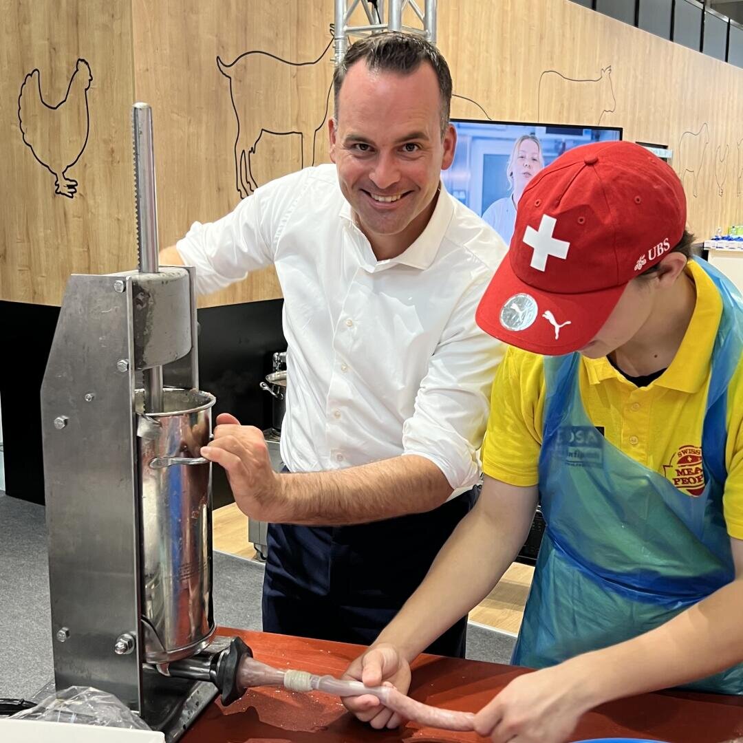 Zwei Personen stellen mit einer Metallmaschine Würste her. Die eine, in einem weißen Hemd, lächelt in die Kamera, während sie die Kurbel dreht. Die andere, die eine rote Mütze und eine Schürze trägt, führt den Wurstdarm in die Düse der Maschine ein.