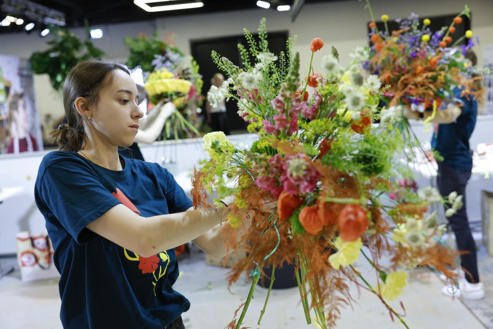 Eine junge Frau arrangiert sorgfältig einen großen, bunten Blumenstrauß in einem hellen Innenraum, umgeben von verschiedenen Blumenauslagen und Werkstattmaterialien.