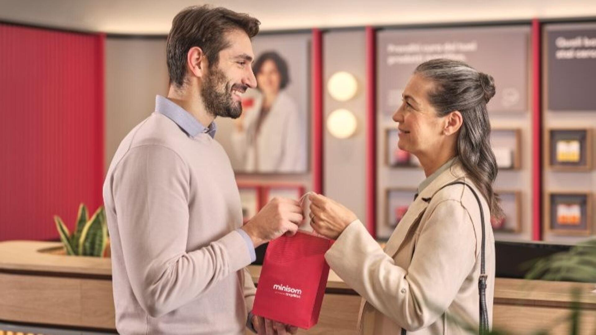 Ein Mann und eine Frau lächeln sich an, während sie gemeinsam eine rote Tasche in einem modernen, hell erleuchteten Geschäft oder Büro mit roten Akzenten und verschwommenen Werbeplakaten im Hintergrund halten.