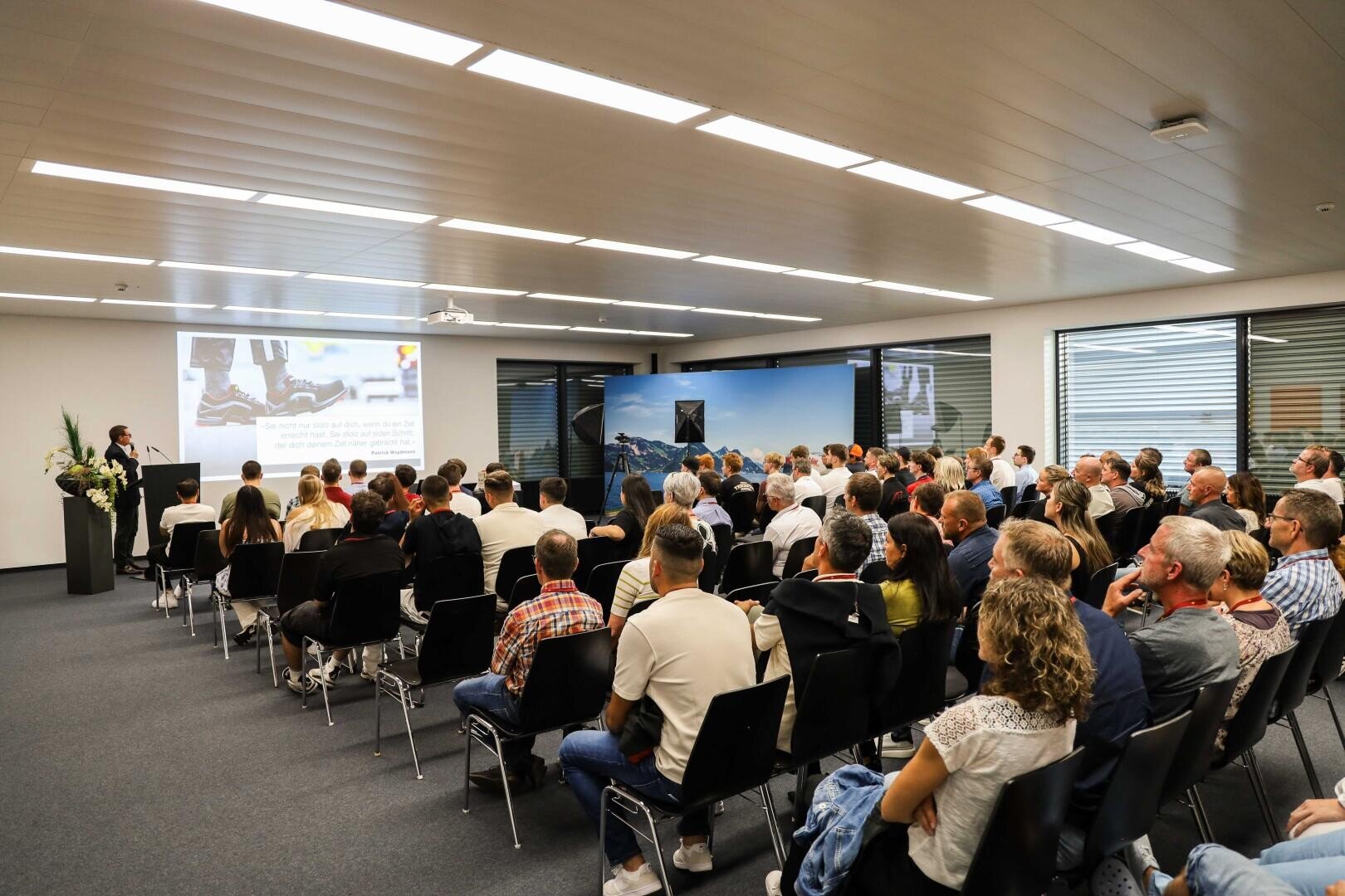 Eine Gruppe von Menschen sitzt in Reihen und hört aufmerksam einem Referenten zu, der vorne im Konferenzraum spricht, während auf einer großen Leinwand eine Präsentation gezeigt wird.