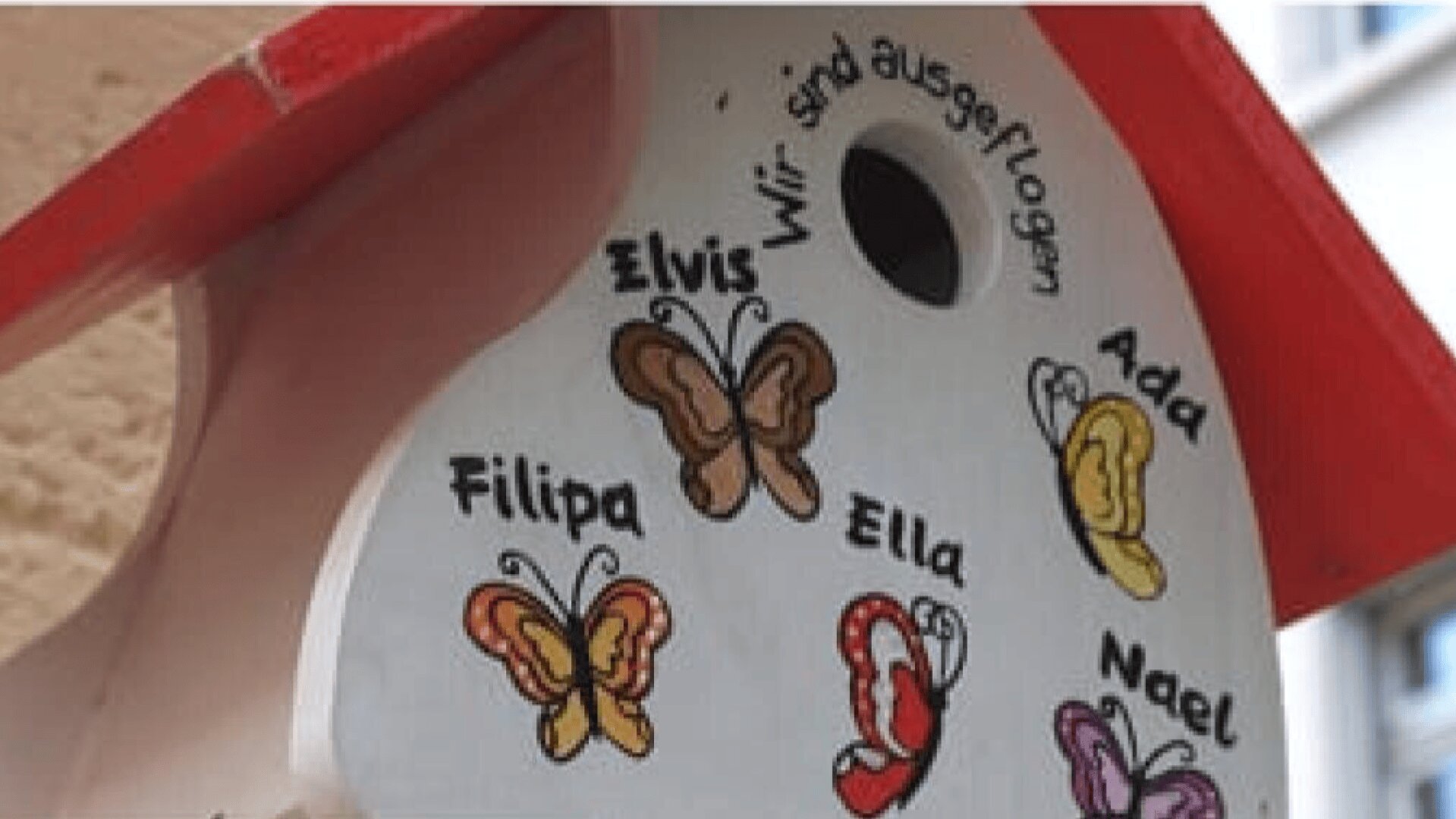 Nahaufnahme eines dekorativen Vogelhauses mit rotem Dach, auf dem Schmetterlinge und handgeschriebene Namen wie Elvis, Filipa, Ella, Ada und Nael sowie der deutsche Satz Wir sind ausgeflogen abgebildet sind.