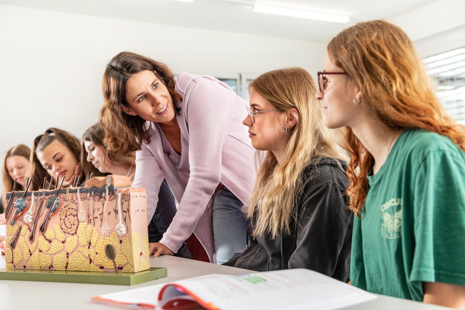 Ein Lehrer beugt sich vor, um mit zwei Schülern in einem Klassenzimmer zu sprechen. Im Hintergrund sind andere Schüler zu sehen, und auf dem Tisch vor ihnen liegt ein großes Modell der menschlichen Haut. Ein aufgeschlagenes Lehrbuch ist ebenfalls zu sehen.
