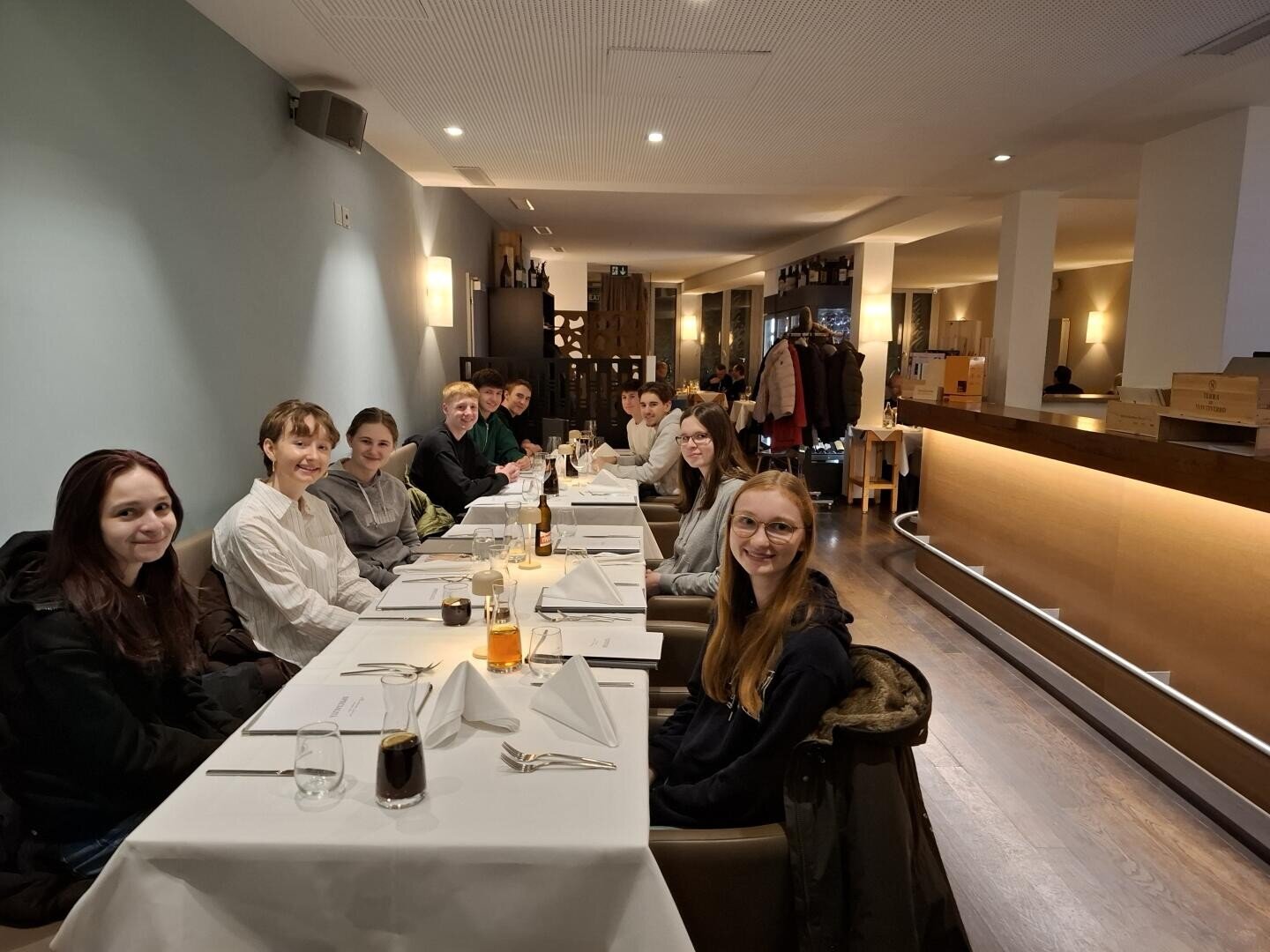Eine Gruppe von zehn Personen sitzt in einem modernen Restaurant an einem langen Tisch und lächelt in die Kamera. Auf dem Tisch stehen Gläser und Getränke. Das Restaurant besticht durch eine warme Beleuchtung und ein minimalistisches Design.