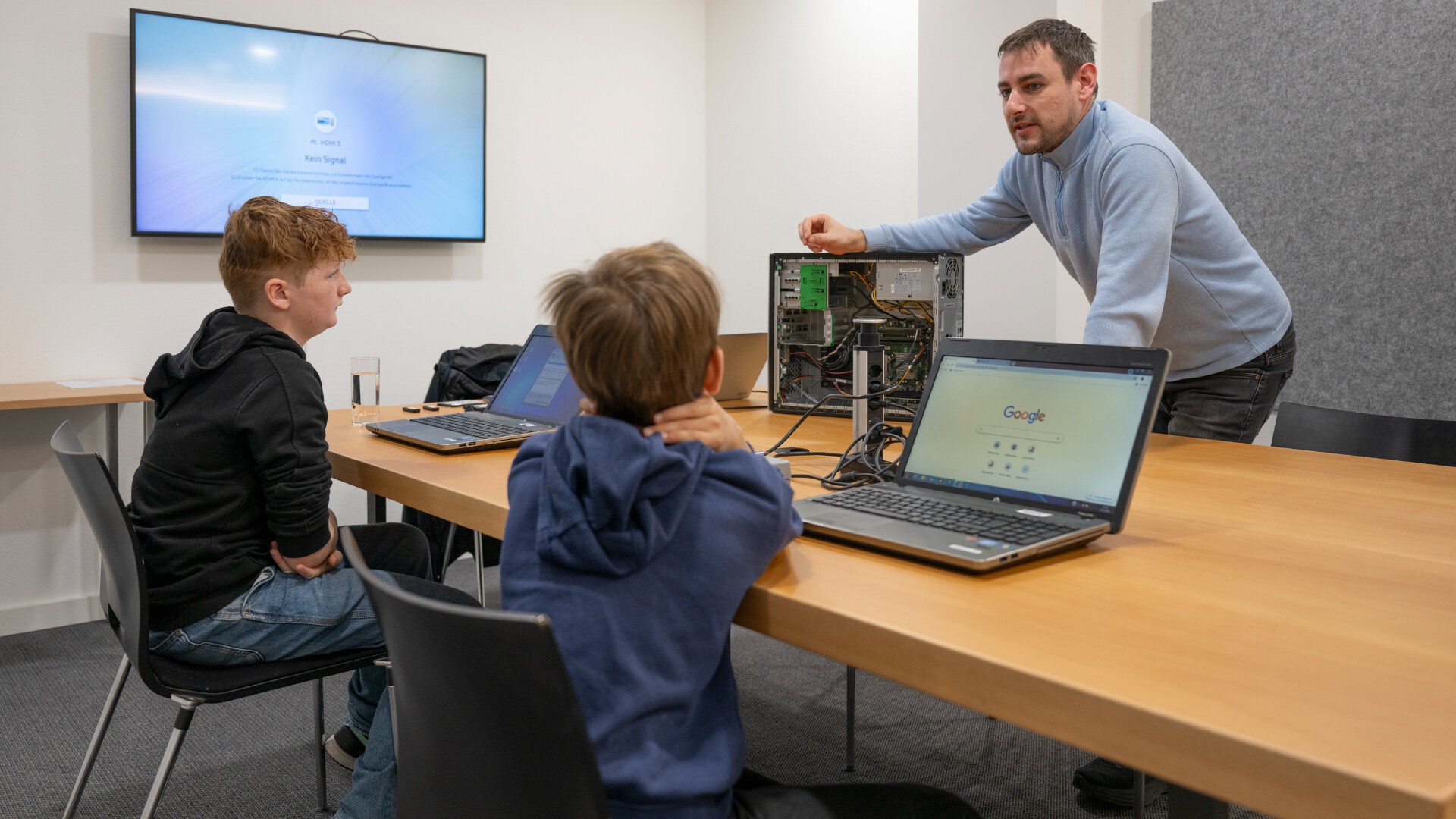 Ein Mann steht neben einem Computerturm und erklärt zwei Jungen, die mit Laptops an einem Tisch sitzen, etwas. Ein Bildschirm an der Wand zeigt ein Anmeldefenster. Die Szene ist in einem Sitzungszimmer.