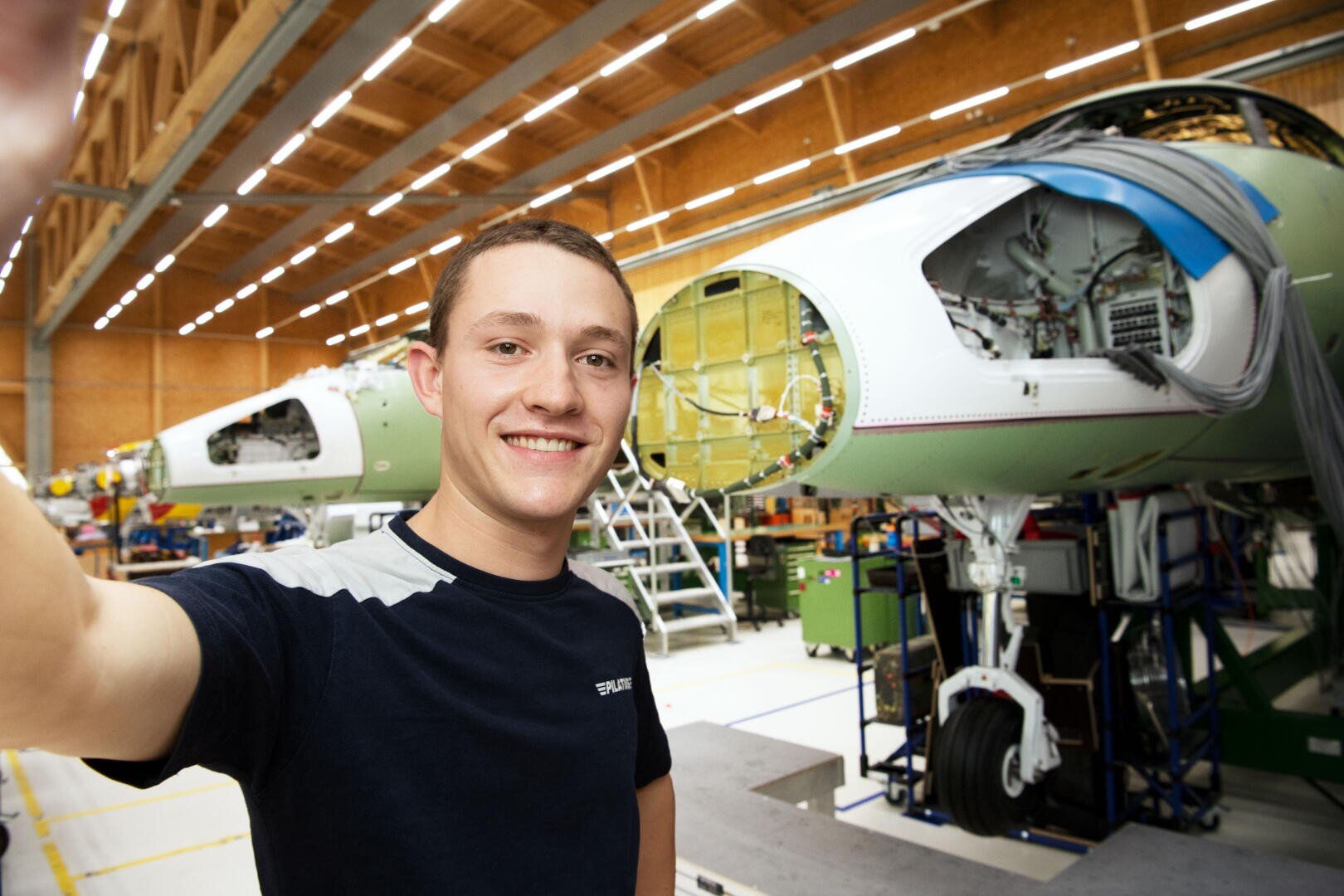 Ein junger Mann lächelt und macht ein Selfie in einer Flugzeugmontagehalle, wobei im hell beleuchteten Hintergrund Flugzeugbauteile und Maschinen zu sehen sind.