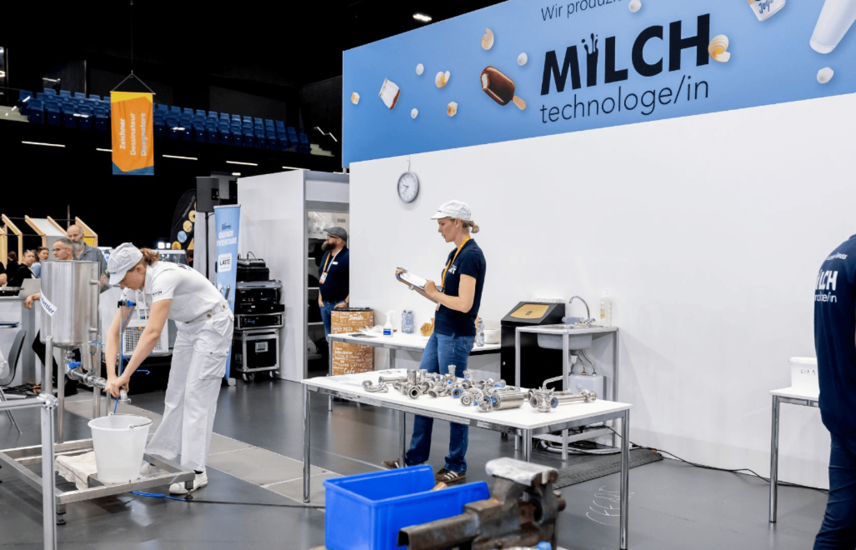 Zwei Personen in weißen Uniformen und Hüten arbeiten an einem Stand mit der Aufschrift Milchtechnologie/in mit Molkereigeräten. Eine Person schreibt auf einem Klemmbrett, während die andere Flüssigkeit in einen Eimer schüttet. Maschinen und Tische sind zu sehen.