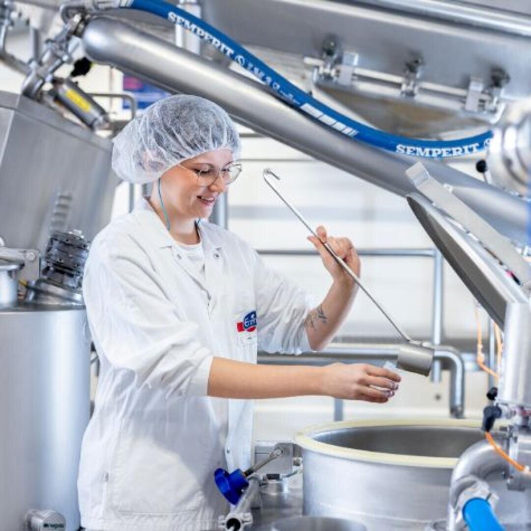 Eine Person in einem weißen Laborkittel, Haarnetz und einer Brille entnimmt mit einem Metallprobenehmer eine Probe aus einem großen Industrietank aus Edelstahl in einem sauberen, modernen Labor oder einer Lebensmittelverarbeitungsanlage.