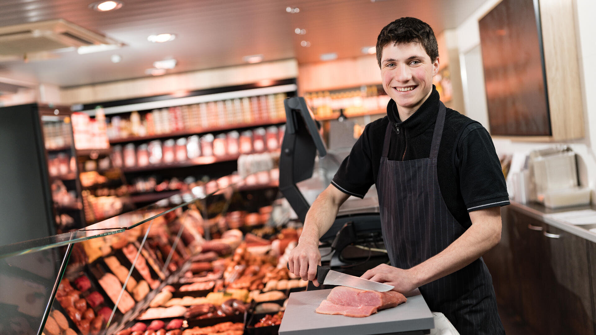 Ein lächelnder Metzger in einer schwarzen Schürze schneidet rohes Fleisch auf einem Schneidebrett hinter einer Fleischtheke mit verschiedenen Fleisch- und Wurstsorten in einem gut beleuchteten Laden.