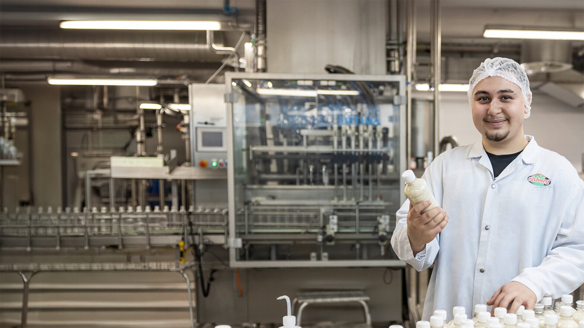 Ein lächelnder Arbeiter in weißem Laborkittel und Haarnetz steht in einer Fabrik und hält eine Produktflasche in der Hand. Flaschen liegen verteilt auf dem Tisch, im Hintergrund sind Maschinen und Förderbänder zu sehen.
