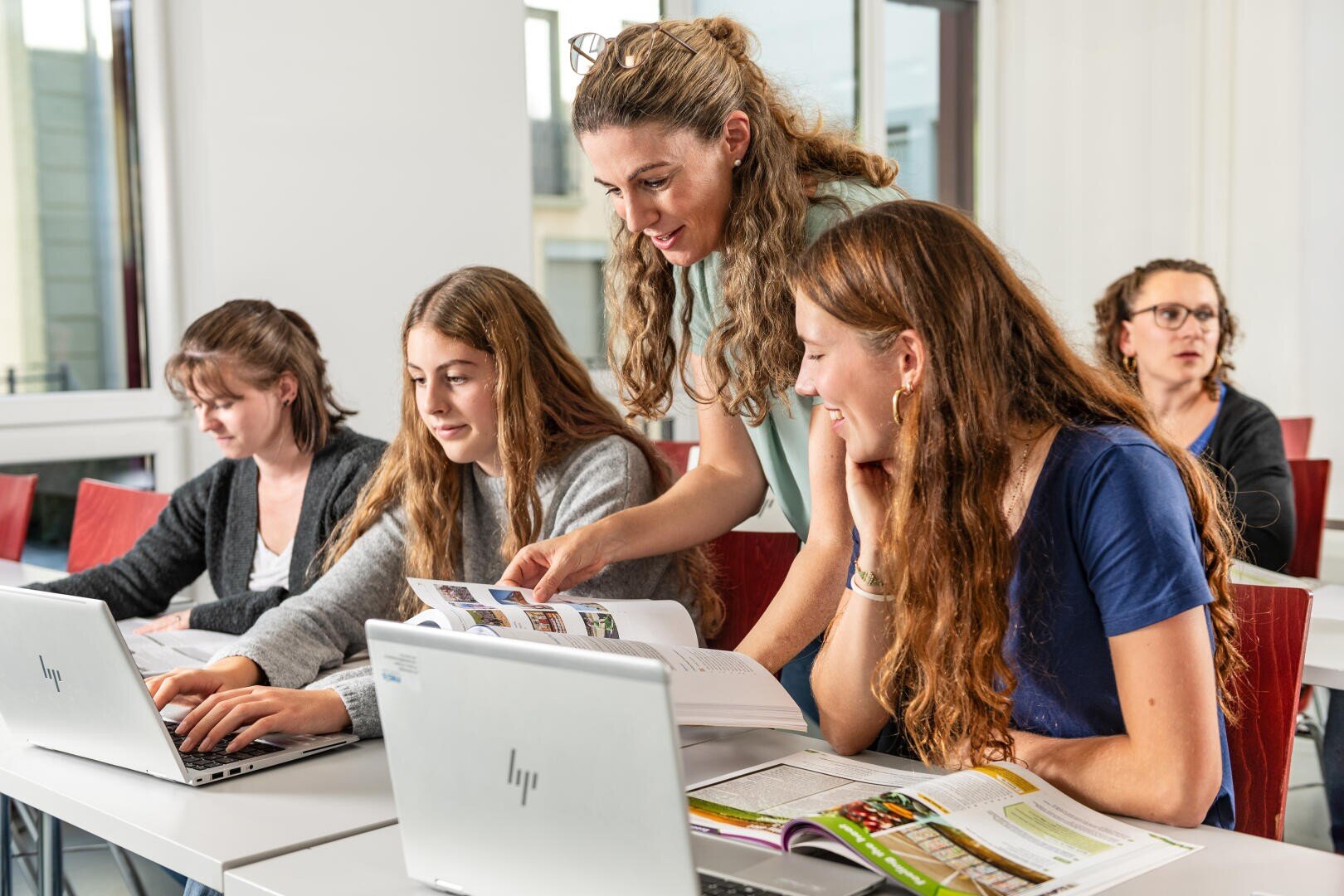 Ein Lehrer hilft zwei Schülern bei ihrer Arbeit, indem er auf ein Lehrbuch zeigt, während die Schüler an Tischen mit Laptops und Papieren sitzen. Zwei weitere Schüler sind im Hintergrund des Klassenzimmers zu sehen.