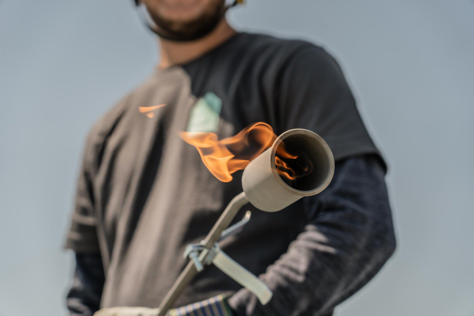 Eine Person in einem dunklen Hemd hält einen Schweißbrenner, aus dessen Düse eine sichtbare orangefarbene Flamme austritt, während sie im Freien vor einem unscharfen Hintergrund steht.