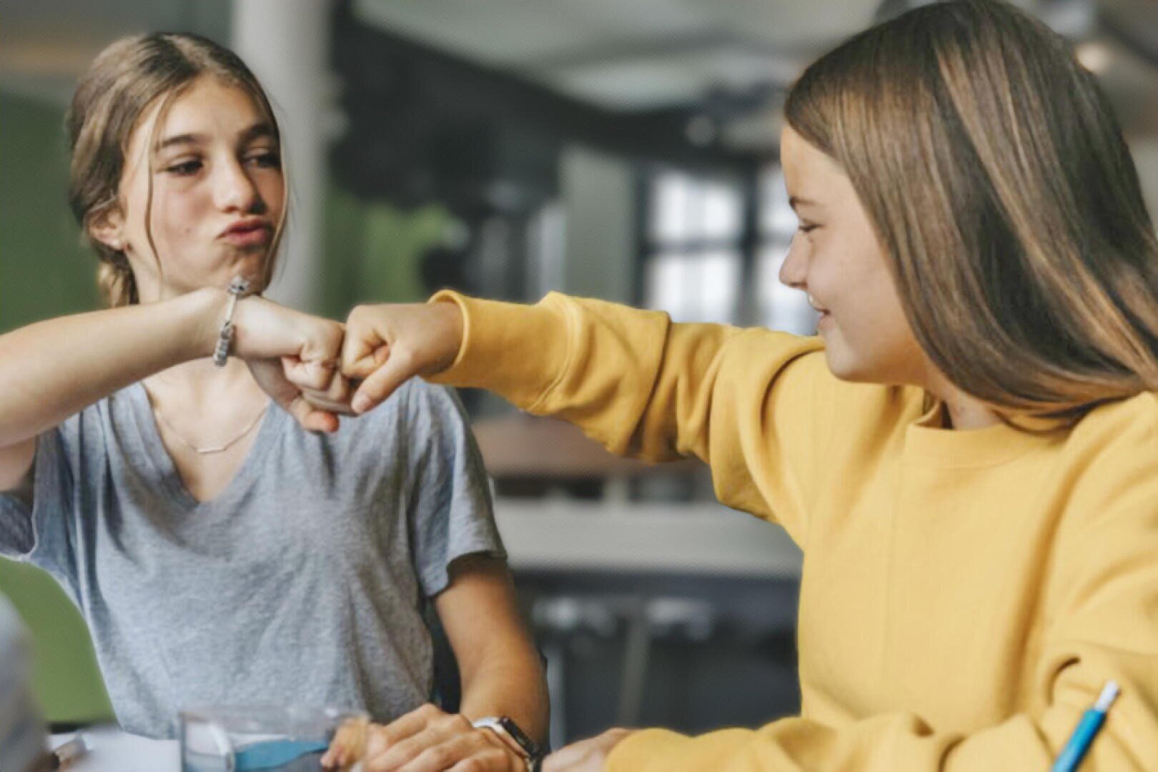 Zwei Mädchen im Teenageralter, die drinnen an einem Tisch sitzen, lächeln und geben sich einen Faustschlag, die eine trägt ein graues Hemd, die andere ein gelbes. Der Hintergrund ist leicht unscharf, was auf eine lockere oder schulische Umgebung hindeutet.