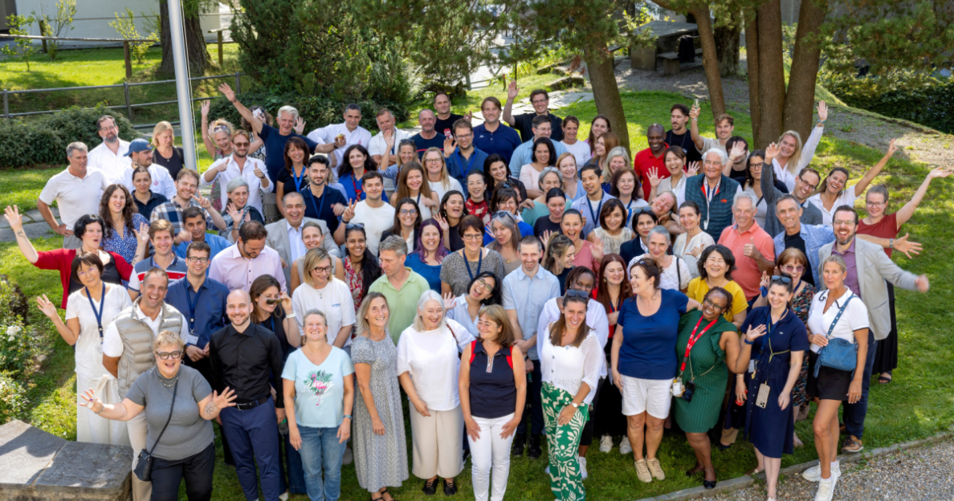 Eine große Gruppe von Menschen unterschiedlichen Alters posiert im Freien auf einer Wiese, lächelt und winkt in die Kamera. Bäume und Grünflächen umgeben sie, und das Wetter scheint sonnig und angenehm.