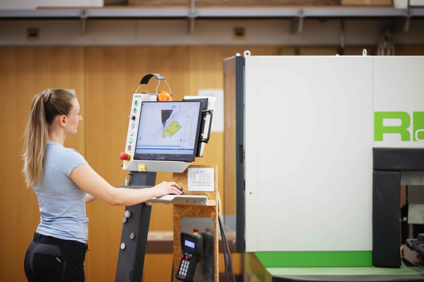 Eine Frau bedient eine CNC-Maschine über ein Bedienfeld mit einem großen Bildschirm, auf dem eine technische Zeichnung in einer Industriehalle angezeigt wird.