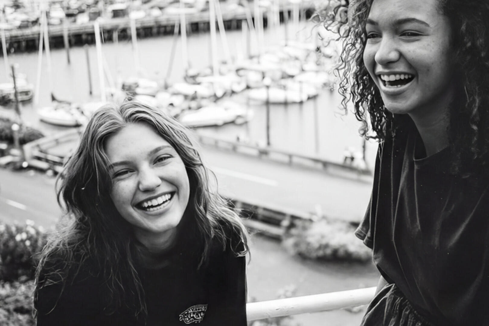 Zwei junge Frauen lächeln und lachen zusammen vor einem Yachthafen mit Booten und Segelbooten im Hintergrund. Das Bild ist in schwarz-weiß gehalten.
