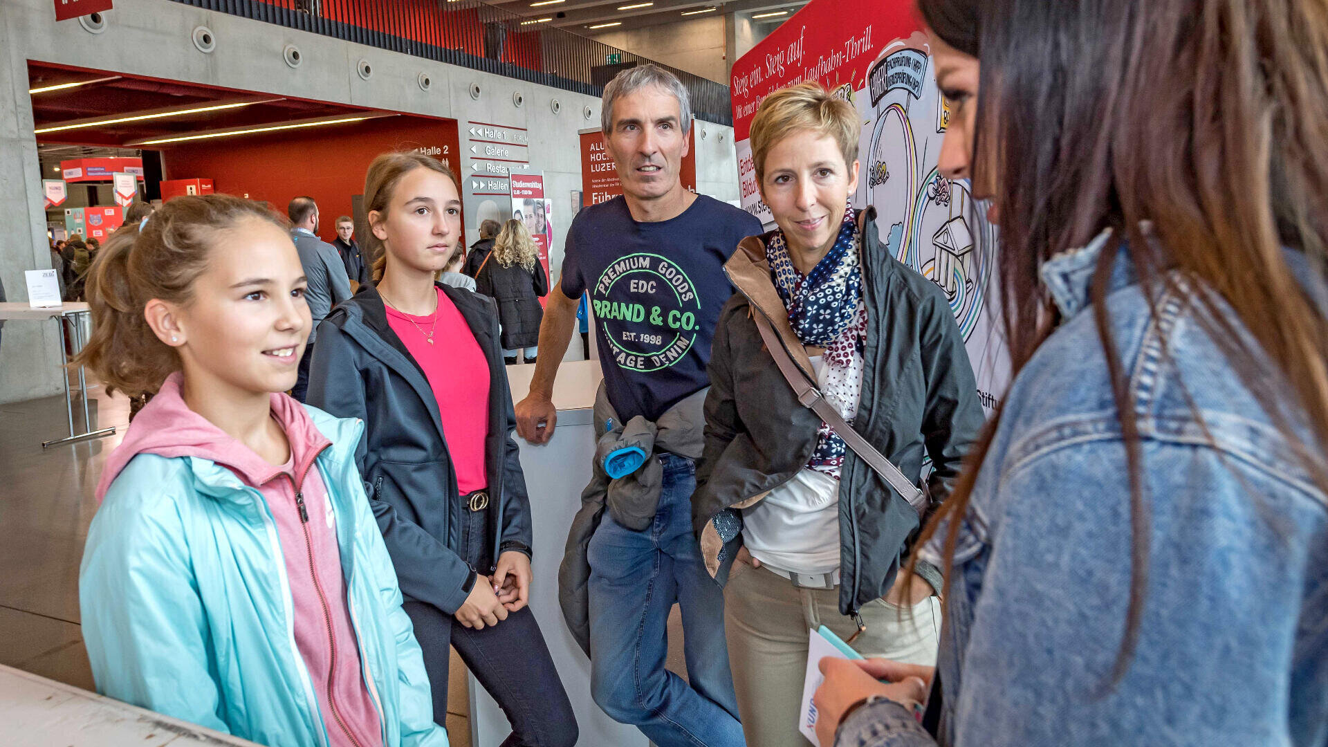 Eine vierköpfige Familie steht bei einer Veranstaltung in einem Saal, lächelt und unterhält sich mit einer Frau mit langen dunklen Haaren, die ihnen gegenübersteht. Die Umgebung ist modern gestaltet, mit roten und grauen Wänden.