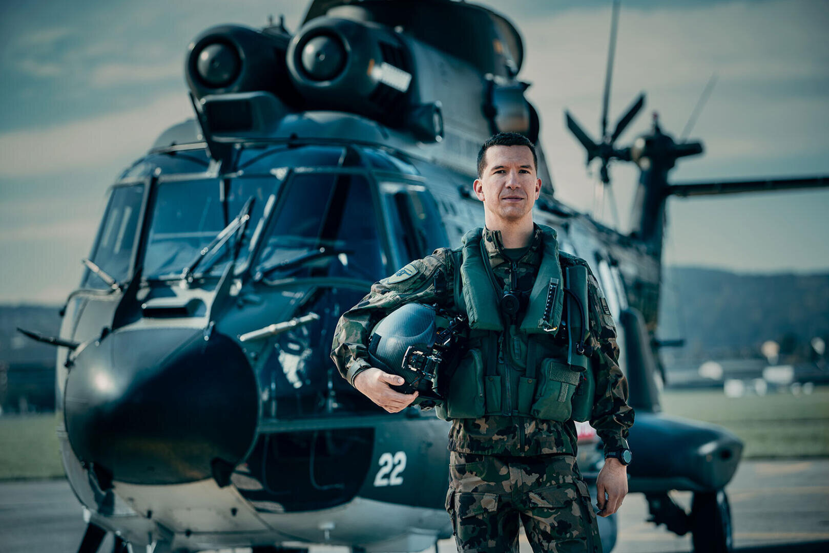 Ein Militärpilot in Tarnuniform und -weste steht vor einem dunklen Hubschrauber und hält einen Helm unter einem Arm auf einem Flugplatz mit einem bewölkten Himmel im Hintergrund.
