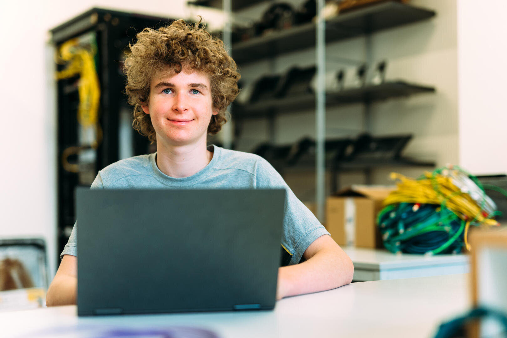 Ein Jugendlicher mit lockigem Haar sitzt an einem Schreibtisch in einem Klassenzimmer oder Labor und lächelt in die Kamera, während er an einem Laptop arbeitet. Im Hintergrund sind Regale mit Geräten und Kabeln zu sehen.