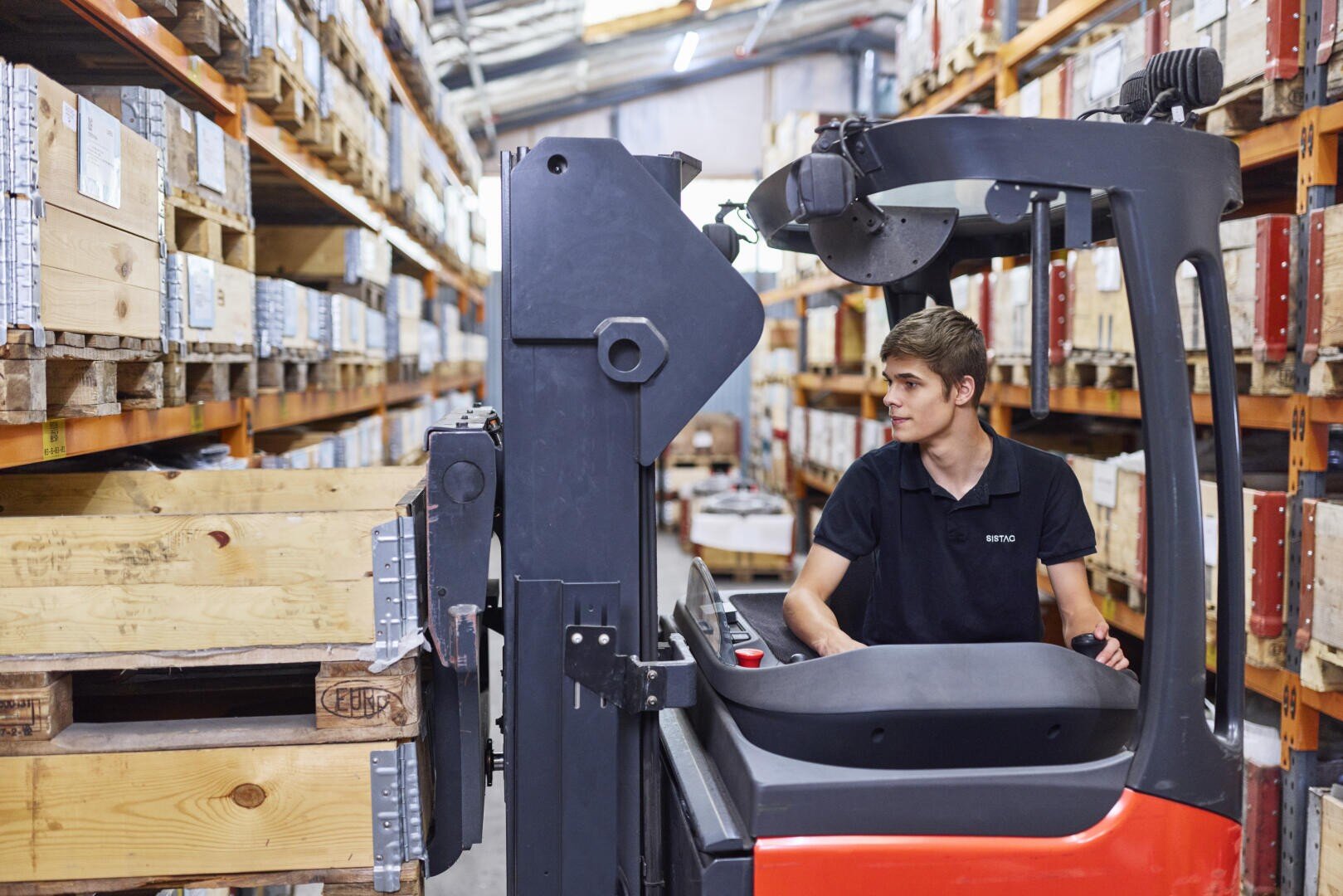 Ein Mann bedient einen Gabelstapler in einem Lagerhaus und bewegt eine große Holzkiste zwischen Regalen, die mit Kisten und Paketen gefüllt sind. Er blickt nach rechts, während er die Maschine steuert.