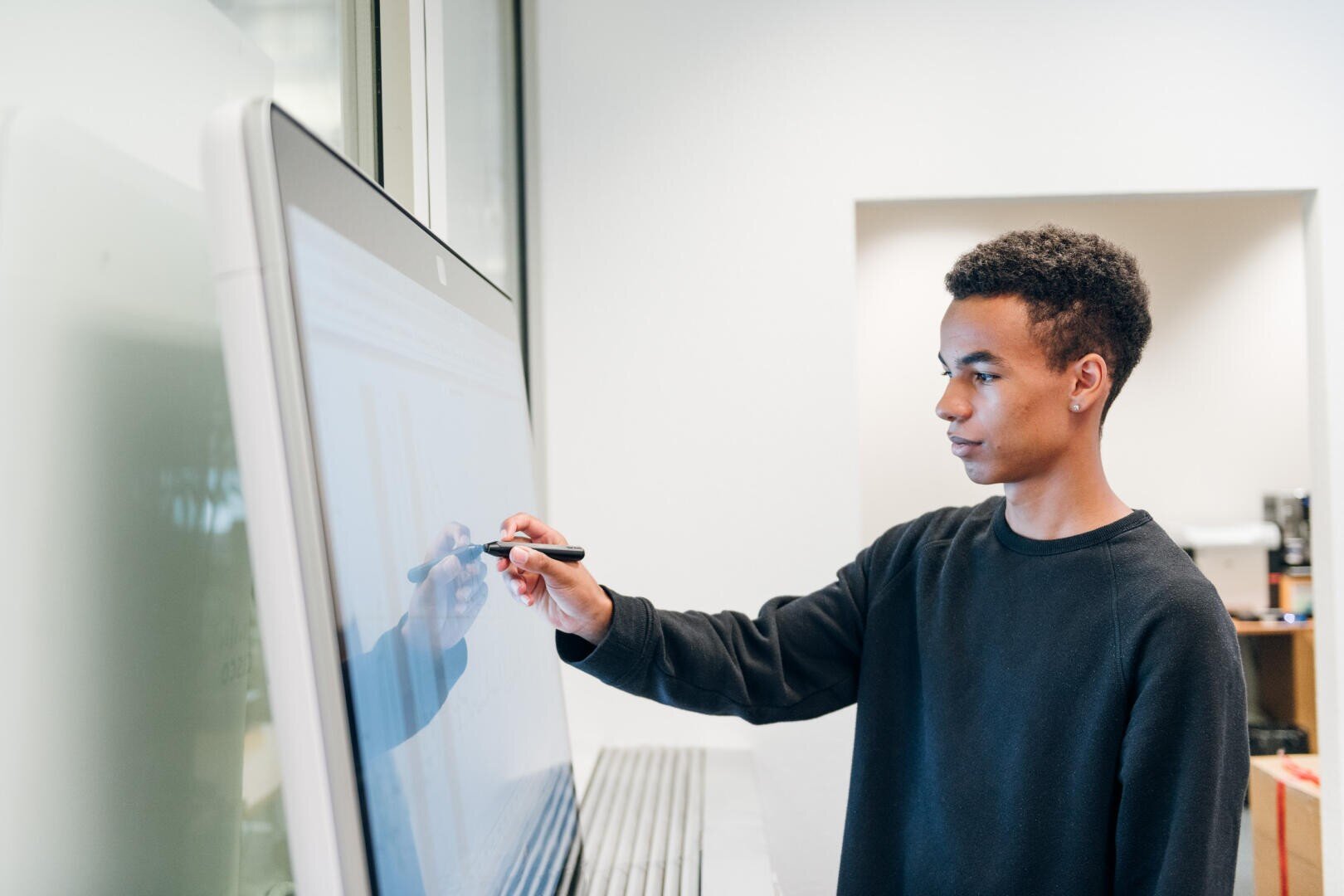 Ein junger Mensch zeichnet oder schreibt mit einem Stift auf einem großen interaktiven Touchscreen in einem hellen, modernen Raum. Sie konzentrieren sich auf den Bildschirm.