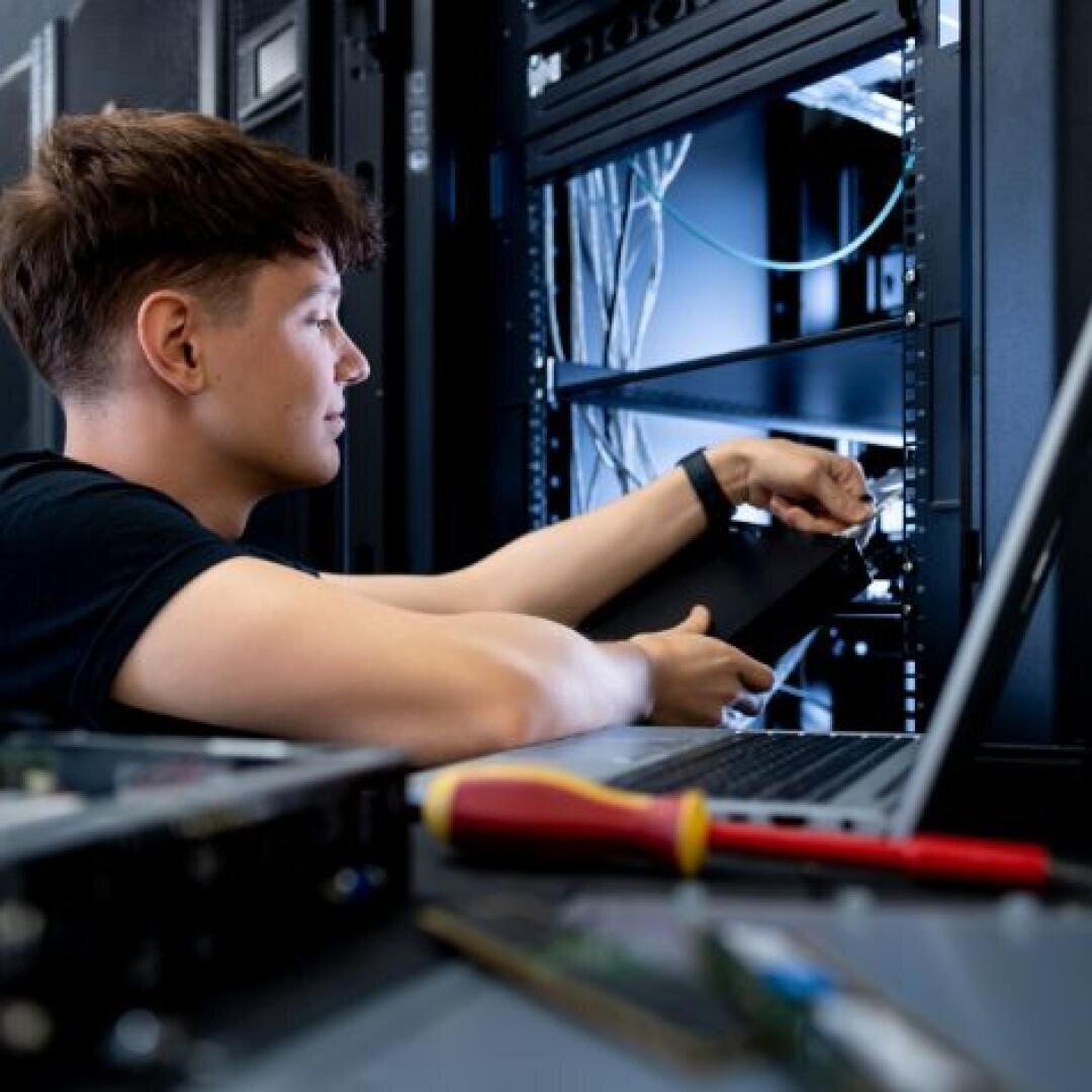 Eine Person arbeitet an einem Serverschrank in einem Rechenzentrum, hält Hardware und benutzt einen Laptop. Auf dem Tisch im Vordergrund sind Werkzeuge zu sehen.