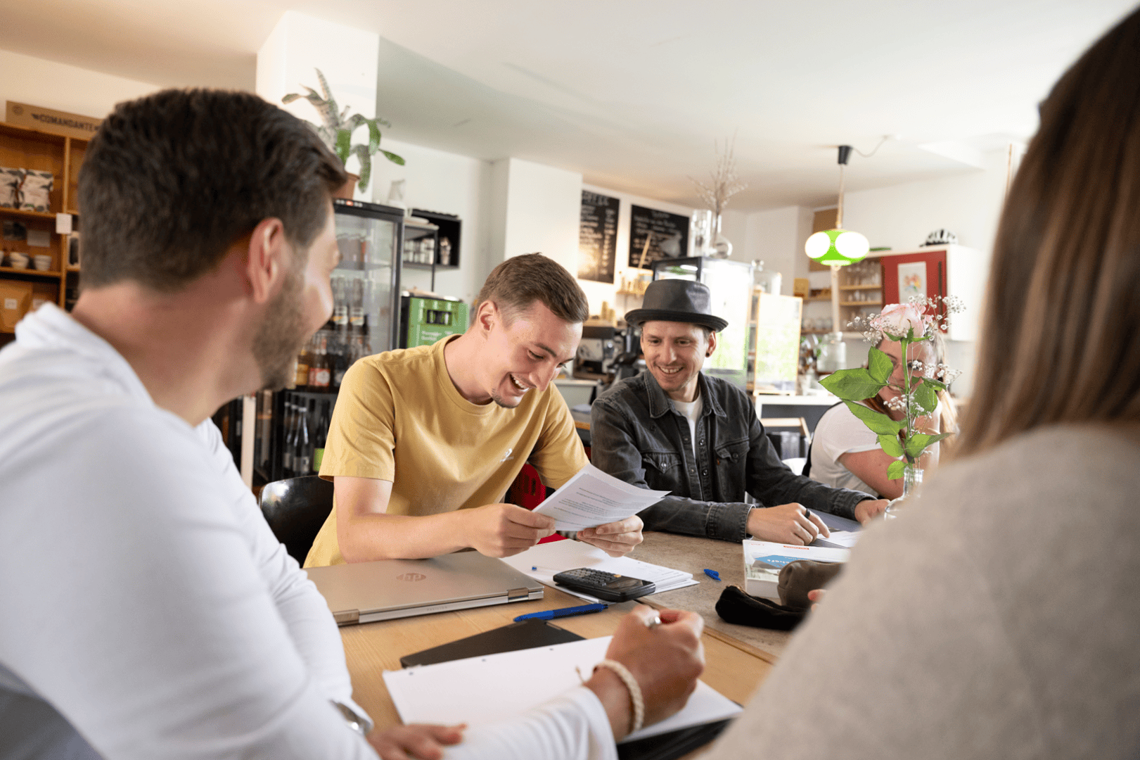 Vier Personen sitzen um einen Tisch in einem hellen, modernen Café, lächeln und unterhalten sich. Ein Mann in einem gelben Hemd liest aus einer Zeitung, während die anderen zuhören und lachen. Auf dem Tisch stehen Laptops, Notebooks und Blumen.