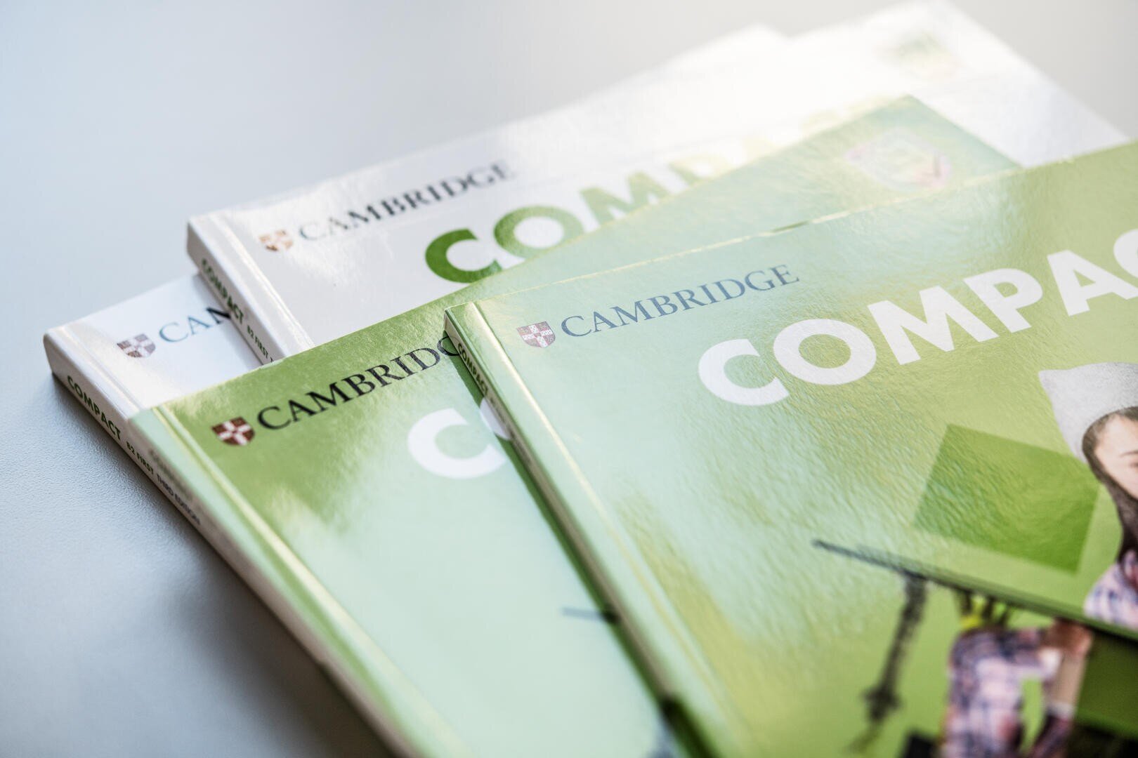Drei Cambridge-Englisch-Lehrbücher mit grünem und weißem Einband, die auf einer hellen Fläche gestapelt sind und das Cambridge-Logo und einen Teil des Titels Compact auf der Vorderseite zeigen.