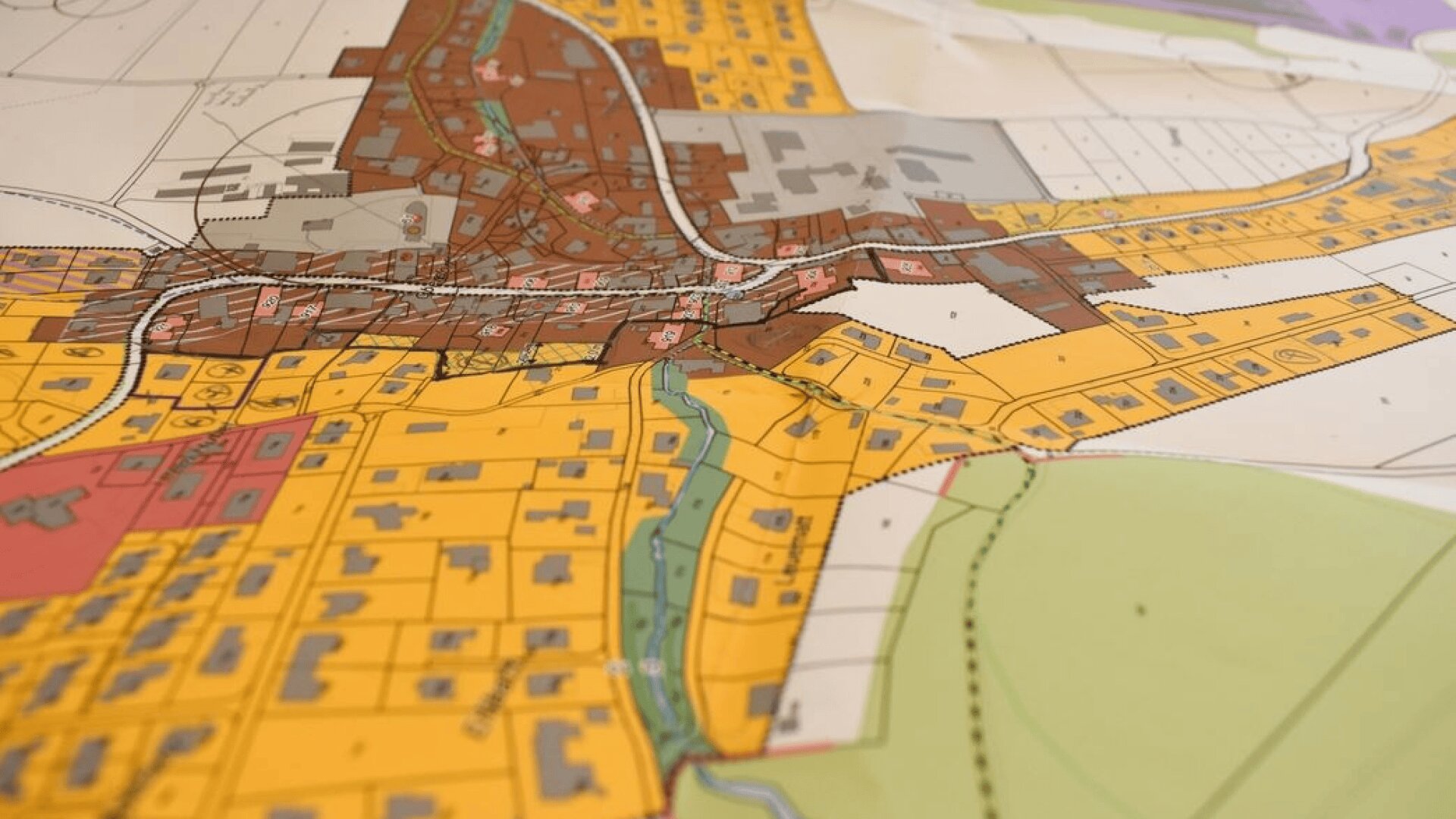 Eine Nahaufnahme einer bunten Stadtplanungskarte mit verschiedenen Zonen, Straßen und Grundstücken in Gelb, Braun, Grün und Grau und einem Fluss in der Mitte.