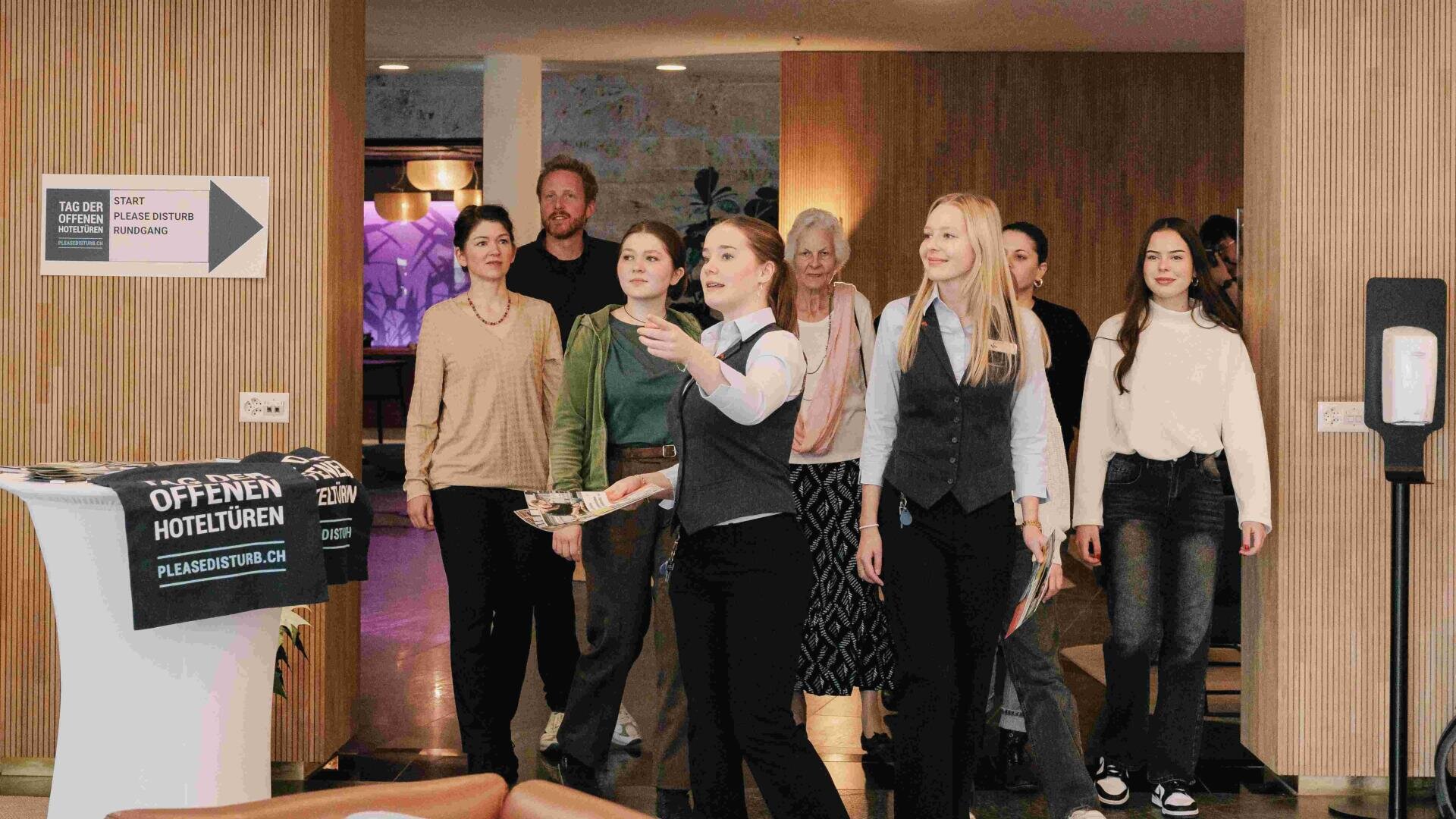 Eine Gruppe von Menschen wird von einer Frau durch die Hotellobby geführt. Sie deutet nach vorne, während die anderen ihr folgen; einige halten Zettel in der Hand. Ein Schild an der Wand weist auf einen Tag der offenen Tür hin.