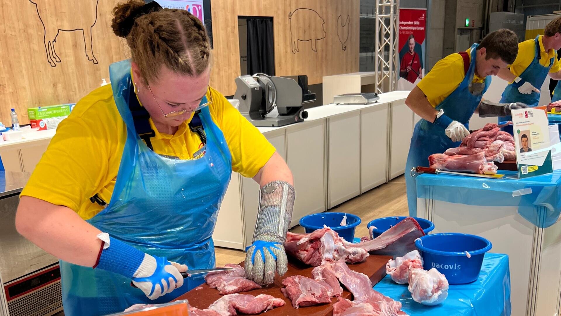 Eine Person, die ein gelbes Hemd, eine blaue Schürze und Schutzhandschuhe trägt, schneidet auf einem Tisch rohes Fleisch, während im Hintergrund eine weitere Person an einem ähnlichen Arbeitsplatz arbeitet.