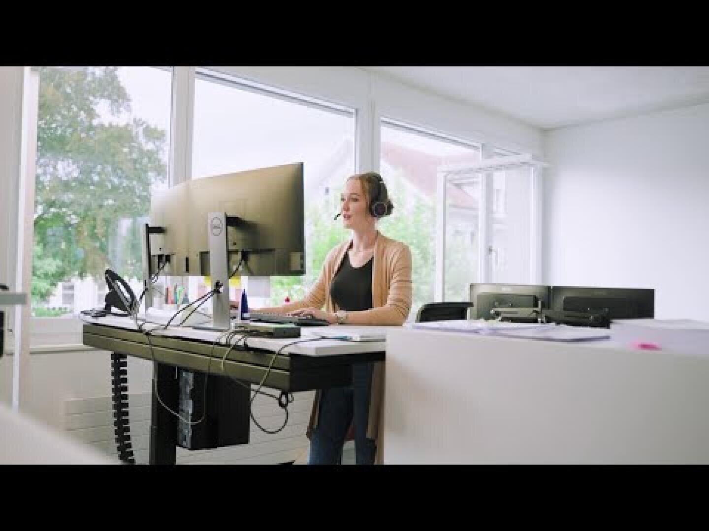 Eine Frau mit Kopfhörern sitzt an einem Schreibtisch mit einem Computer in einem hellen, modernen Büro mit großen Fenstern und Blick ins Grüne. Sie lächelt und scheint an einem Videoanruf oder einer Onlinebesprechung teilzunehmen.