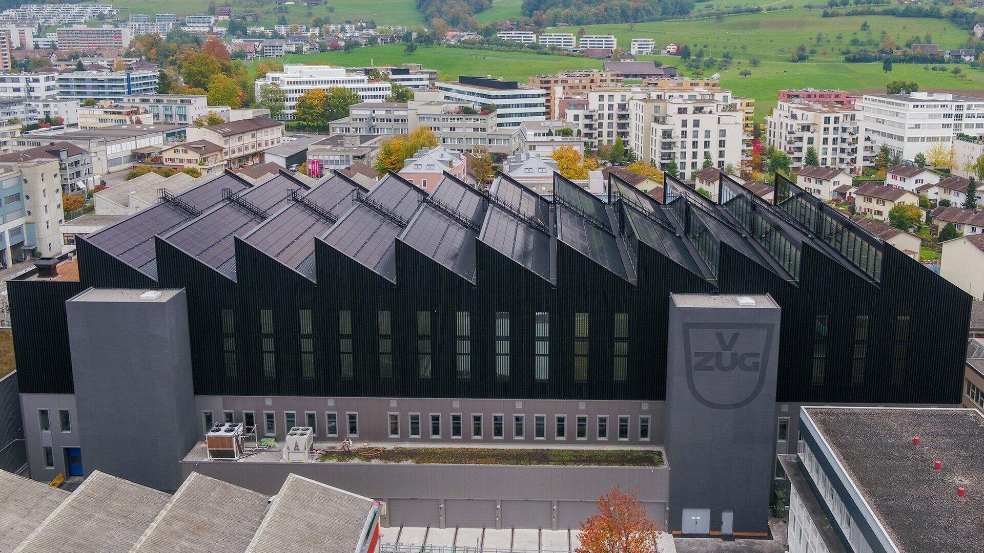 Ein modernes, schwarzes Industriegebäude mit einem Sägezahndach sticht zwischen Wohn- und Geschäftshäusern hervor. An der Fassade ist das Logo V-ZUG zu sehen. Im Hintergrund sind grüne Hügel und Bäume zu sehen.