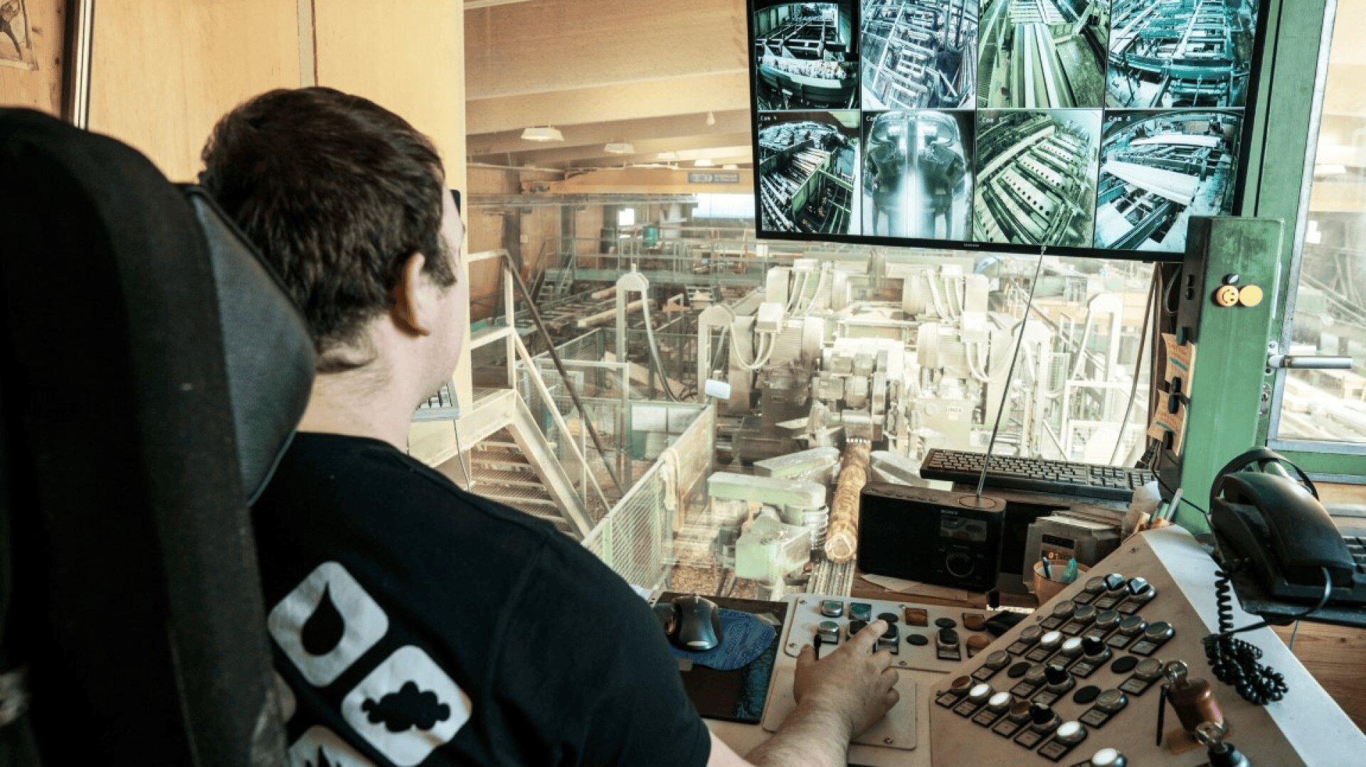 Eine Person sitzt an einer Schalttafel mit Blick auf eine Holzverarbeitungsanlage und überwacht mehrere Überwachungsbildschirme, die verschiedene Ansichten von Maschinen und Stämmen in der Industrieanlage zeigen.