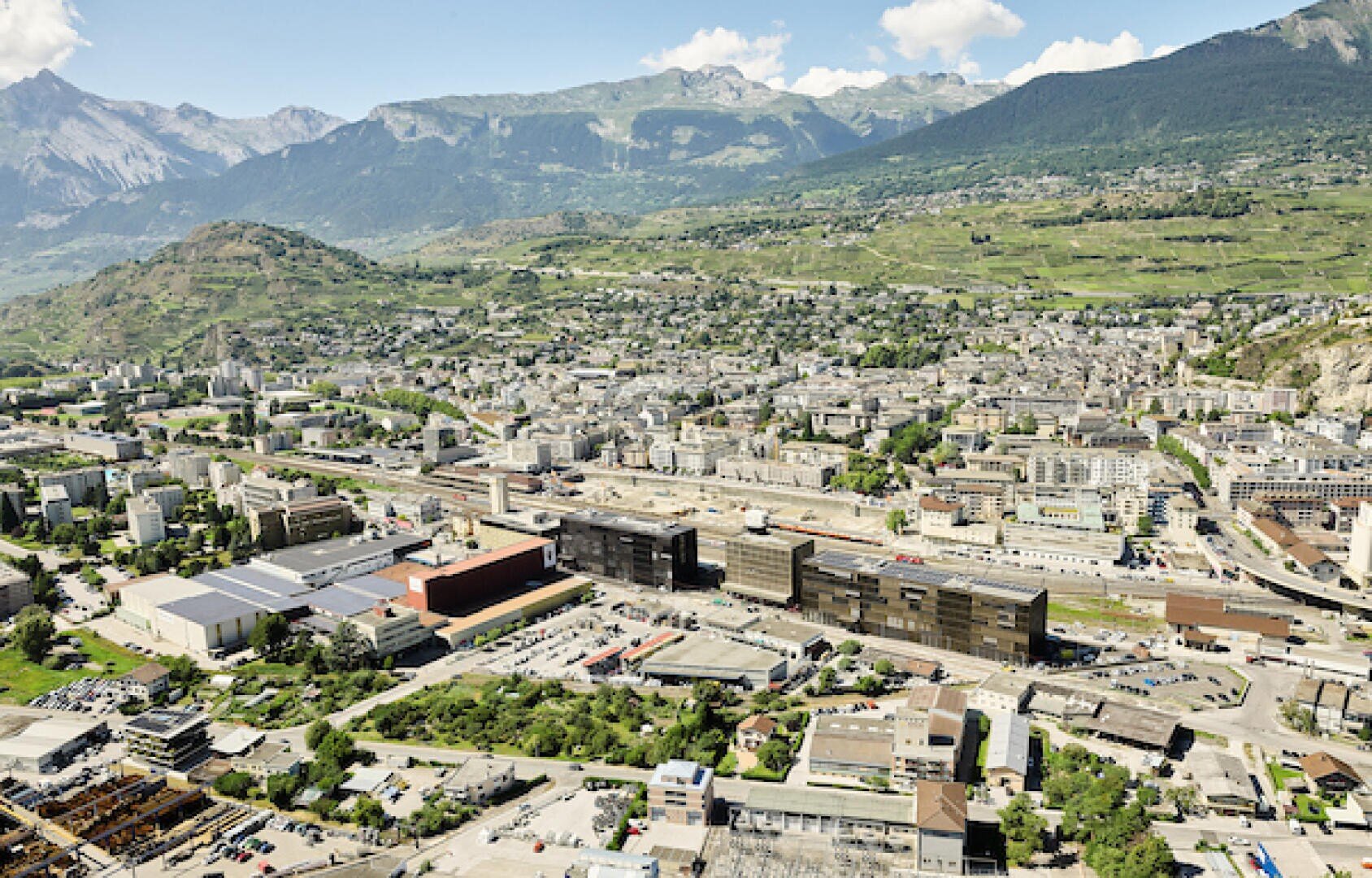 Luftaufnahme einer Schweizer Stadt in einem grünen Tal, mit modernen und älteren Gebäuden, umgeben von sanften Hügeln und Bergen unter einem strahlend blauen Himmel mit vereinzelten Wolken.