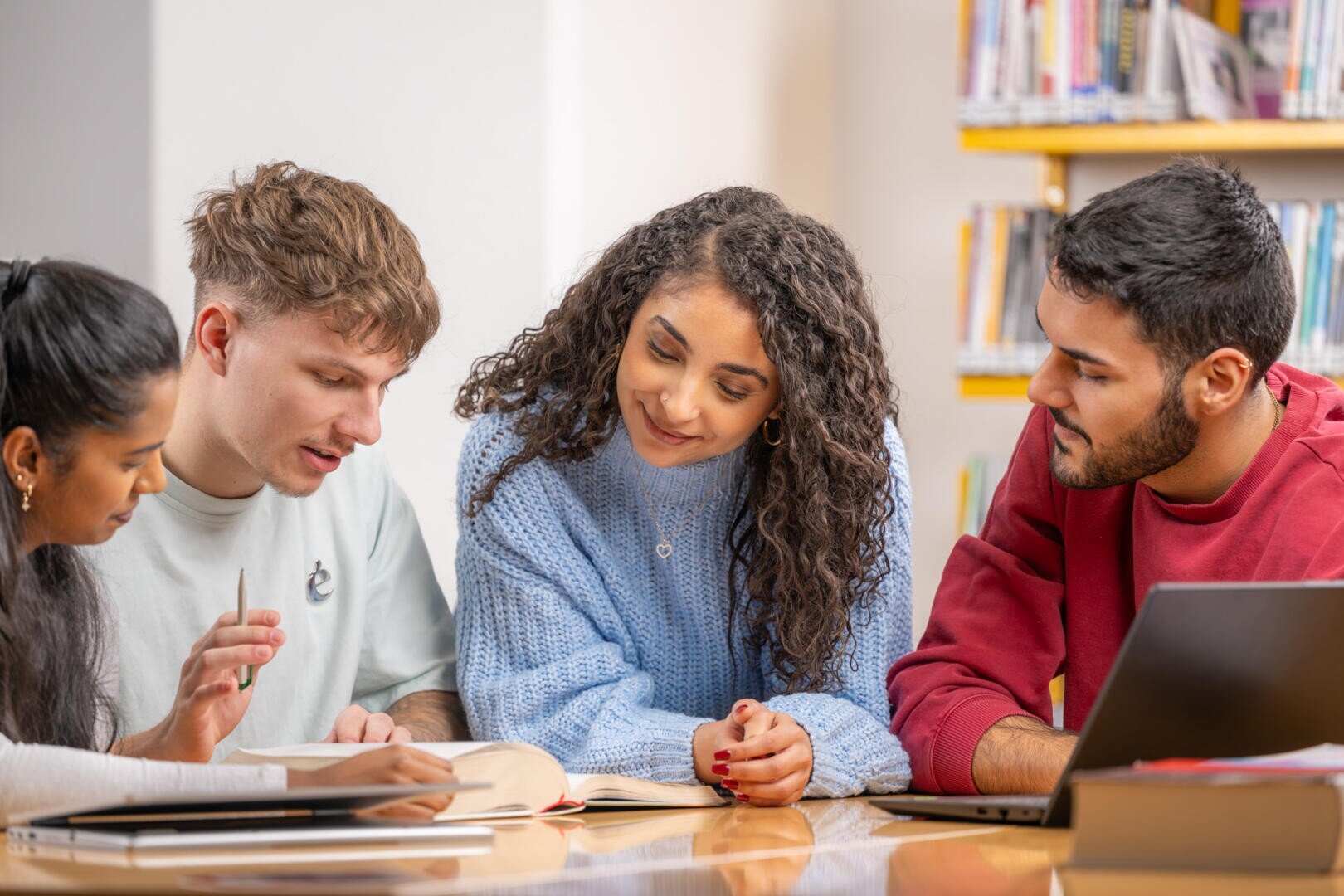 Vier Studenten sitzen an einem Tisch in einer Bibliothek und arbeiten über Büchern und einem Laptop zusammen. Sie scheinen in eine Diskussion vertieft zu sein, wobei eine Person auf ein aufgeschlagenes Buch zeigt, während die anderen aufmerksam zuhören.
