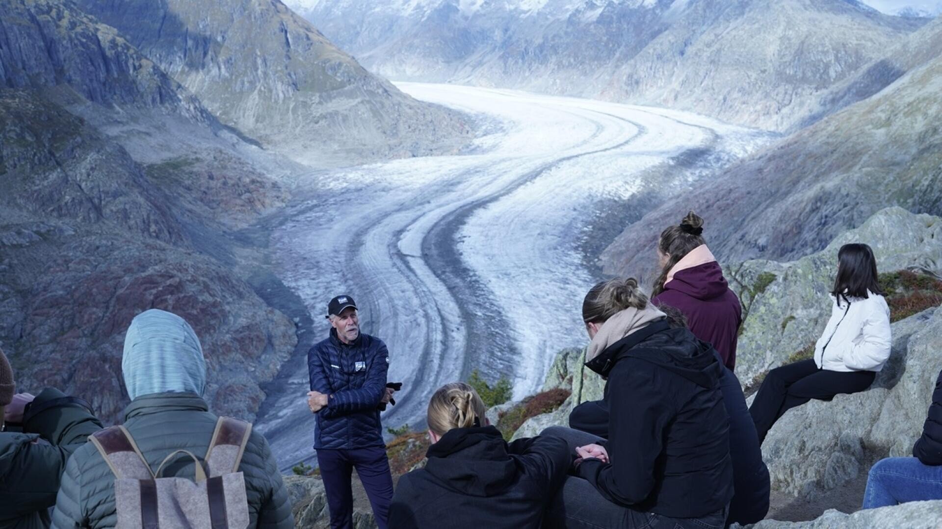 Eine Gruppe von Tourismusstudenten sitzt auf den Felsen mit Blick auf den Aletschgletscher und nimmt an einem Vortrag über die Bedeutung der Gletscher und der Alpenwelt für den Tourismus teil.