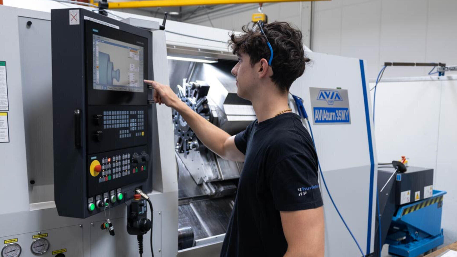 Eine Person bedient eine CNC-Maschine in einer Werkstatt und verwendet das Touchscreen-Bedienfeld, um Einstellungen vorzunehmen. Im Hintergrund sind Industrieanlagen und Werkzeuge zu sehen.