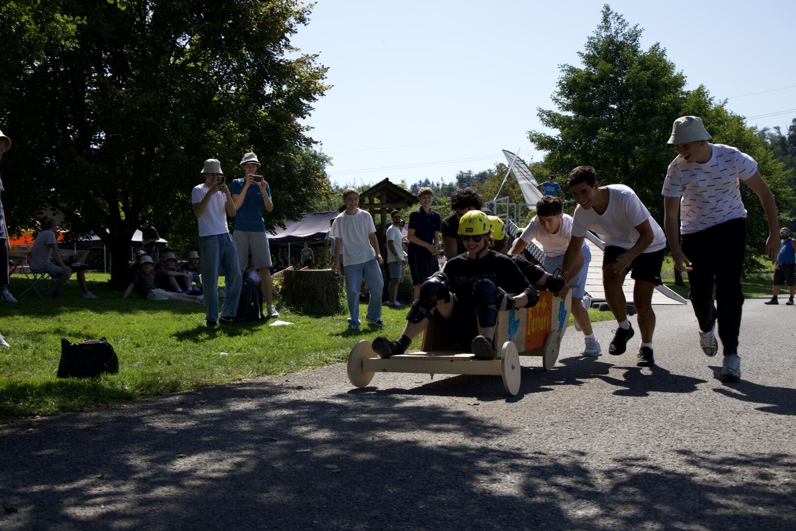 Eine Gruppe junger Leute schiebt ein selbstgebautes Seifenkistenauto aus Holz mit einem behelmten Fahrer einen gepflasterten Weg hinunter, während Zuschauer an einem sonnigen Tag in einem Park zusehen und Fotos machen.