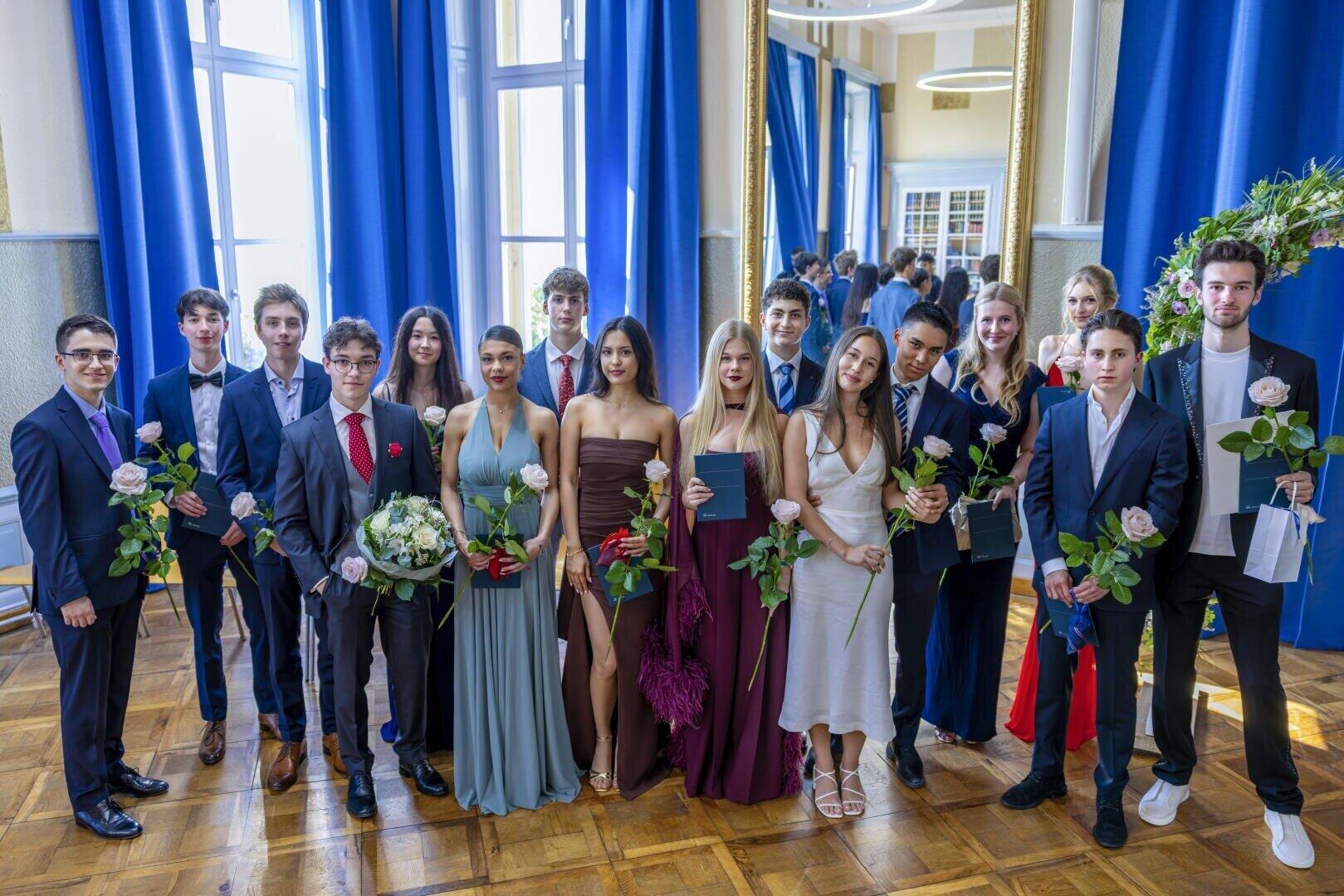In einem hellen Raum mit blauen Vorhängen und großen Fenstern posiert eine Gruppe junger Erwachsener in förmlicher Kleidung mit Urkunden und Blumen in der Hand.