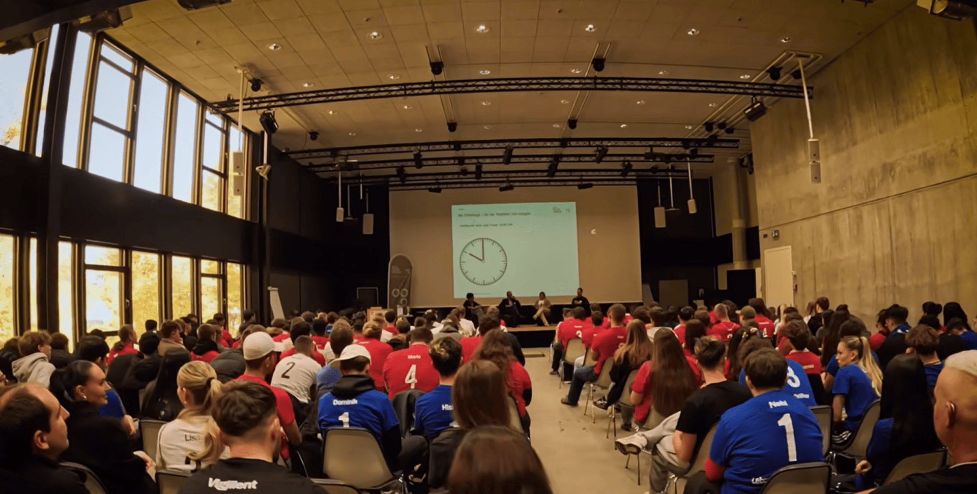 Eine große Gruppe von Menschen, viele in roten oder blauen Hemden, sitzt in einem großen Saal mit hohen Decken vor einer Bühne. Eine Projektion zeigt eine Uhr auf einer Leinwand an der Stirnseite des Raums.