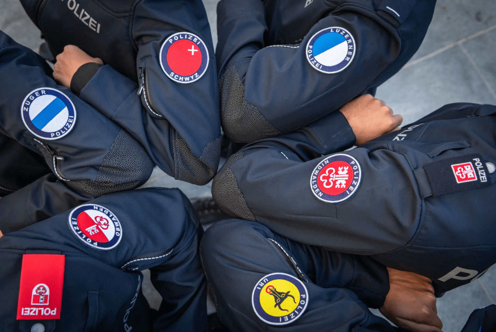 Fünf Polizeibeamte in dunklen Uniformen stehen mit verschränkten Armen im Kreis und tragen verschiedene Ärmelabzeichen der Schweizer Kantonspolizei.