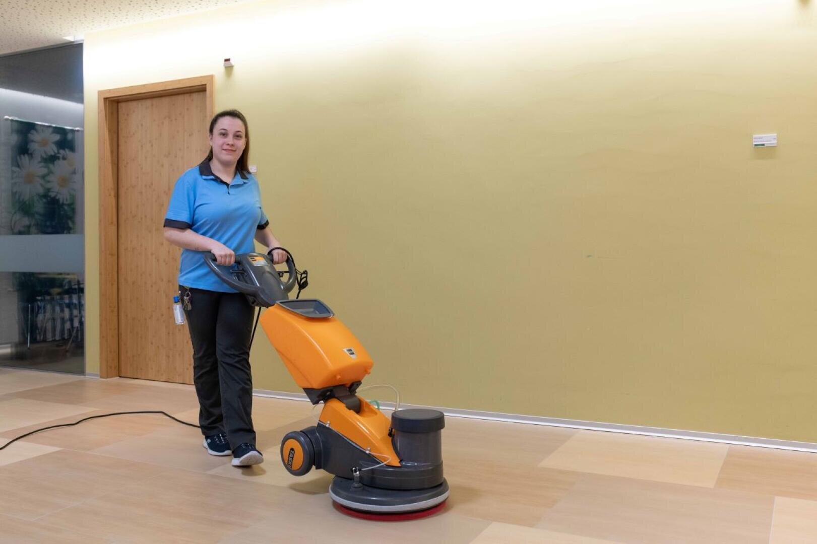 Eine Frau in blauer Uniform bedient eine orangefarbene Bodenreinigungsmaschine auf einem hellen Holzboden in einem hell erleuchteten Raum mit einer gelben Wand und einer geschlossenen Holztür.