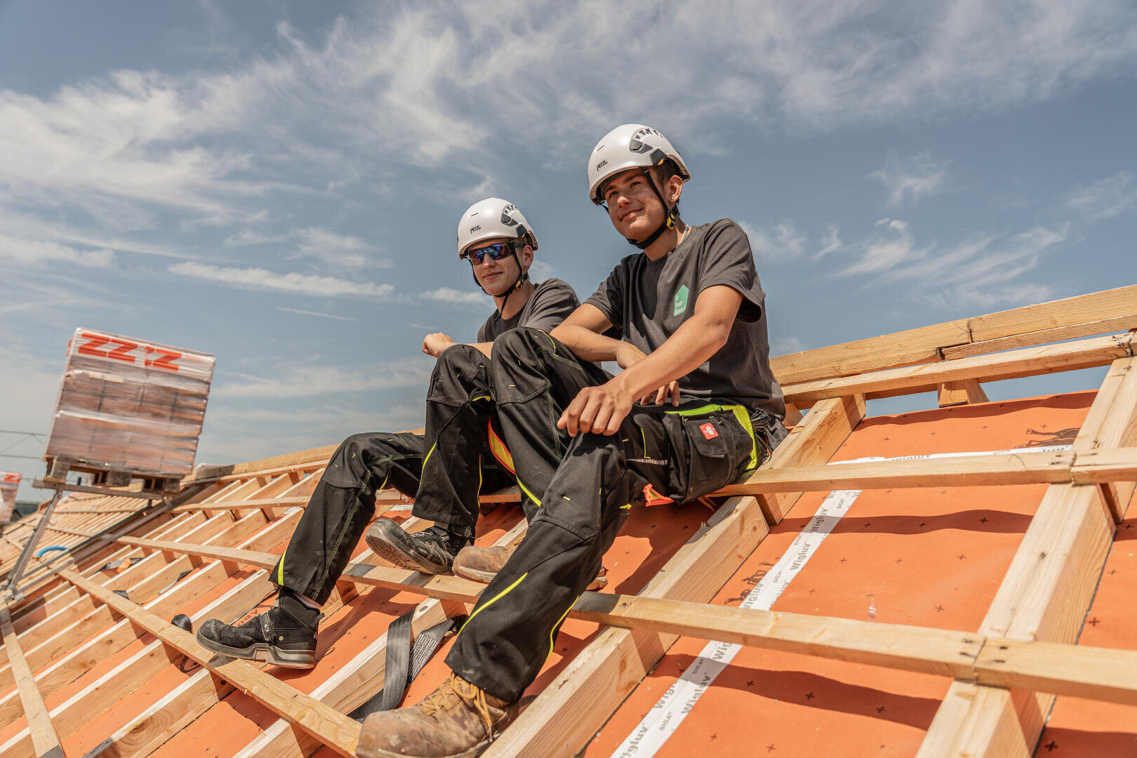 Zwei Bauarbeiter mit Helmen und Arbeitskleidung sitzen auf einem hölzernen Dachstuhl unter einem blauen Himmel und lächeln in die Kamera. In der Nähe sind Baumaterialien und Werkzeuge zu sehen.