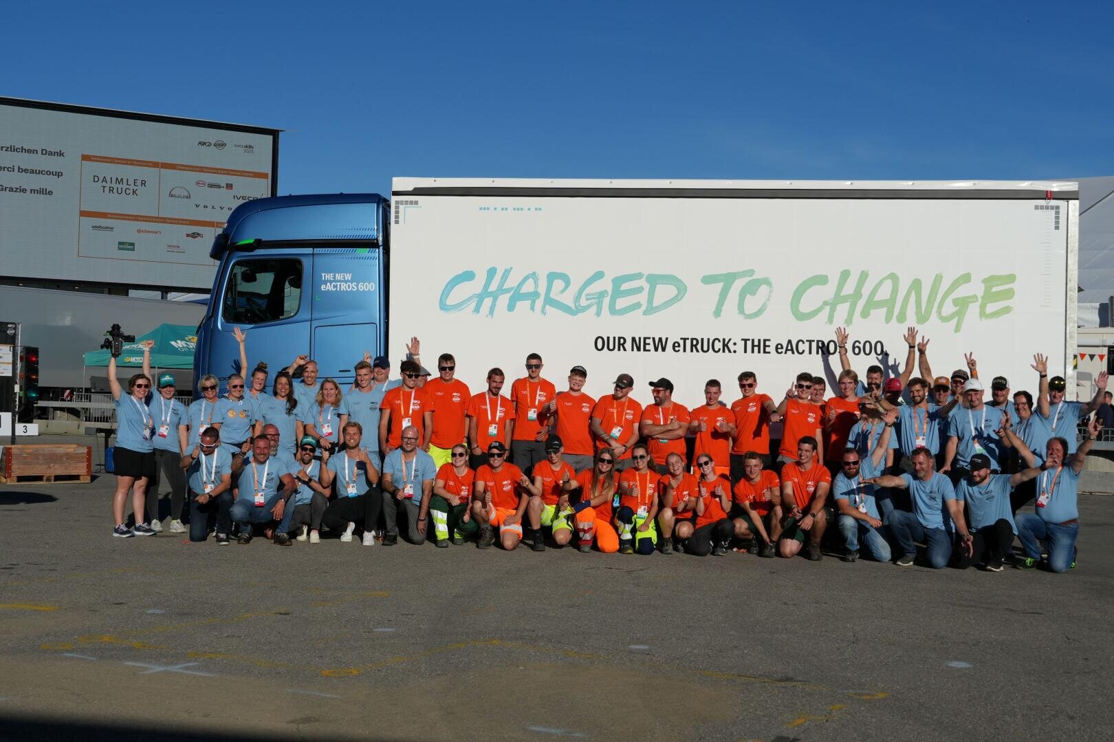 Eine große Gruppe von Menschen in blauen und orangefarbenen T-Shirts posiert vor einem Lkw, auf dessen Seite die Aufschriften „CHARGED TO CHANGE“ und „UNSER NEUER eTRUCK: DER eACTROS 600“ zu lesen sind. Die Szene spielt im Freien an einem sonnigen Tag.